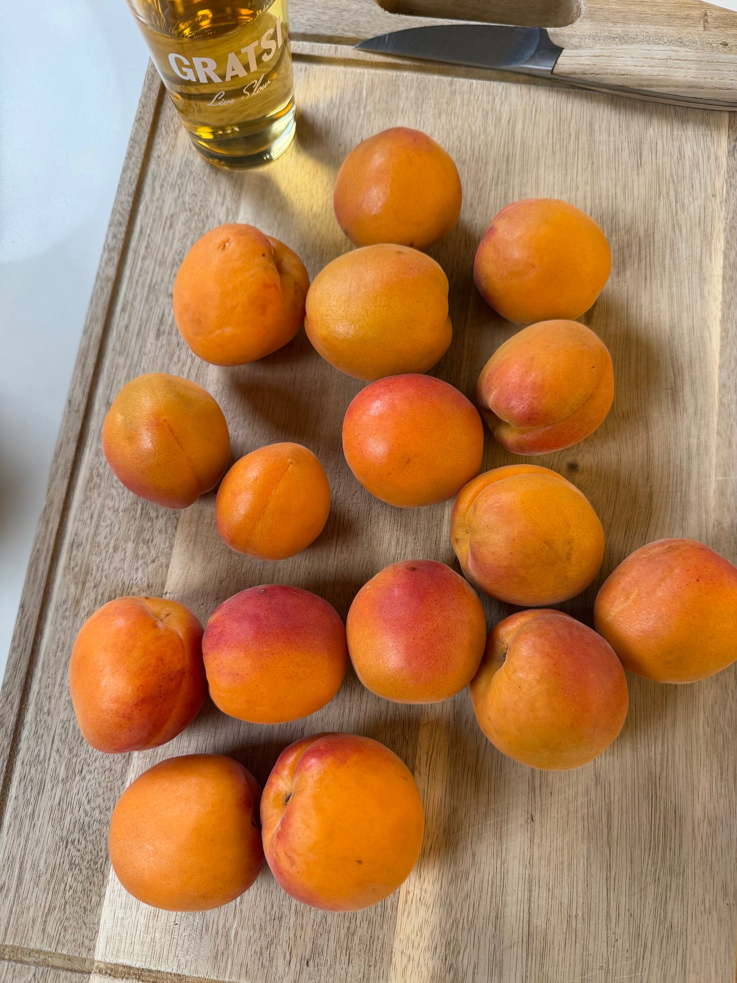A group of 15 ripe apricots is arranged on a wooden cutting board next to a glass of amber-colored liquid.