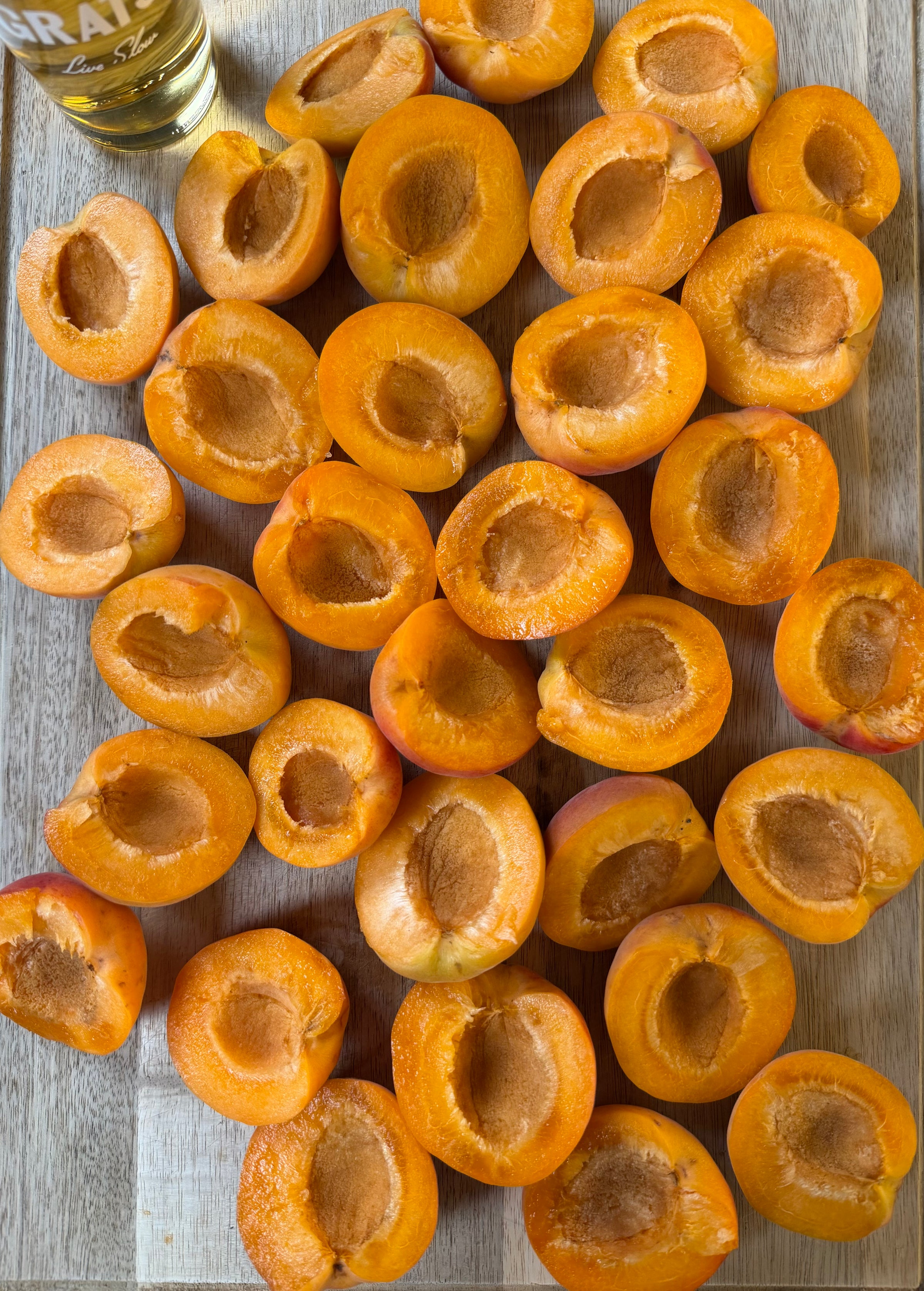 Halved apricots with pits removed are arranged on a wooden surface. A small part of a glass with light-colored liquid is visible in the upper left corner.