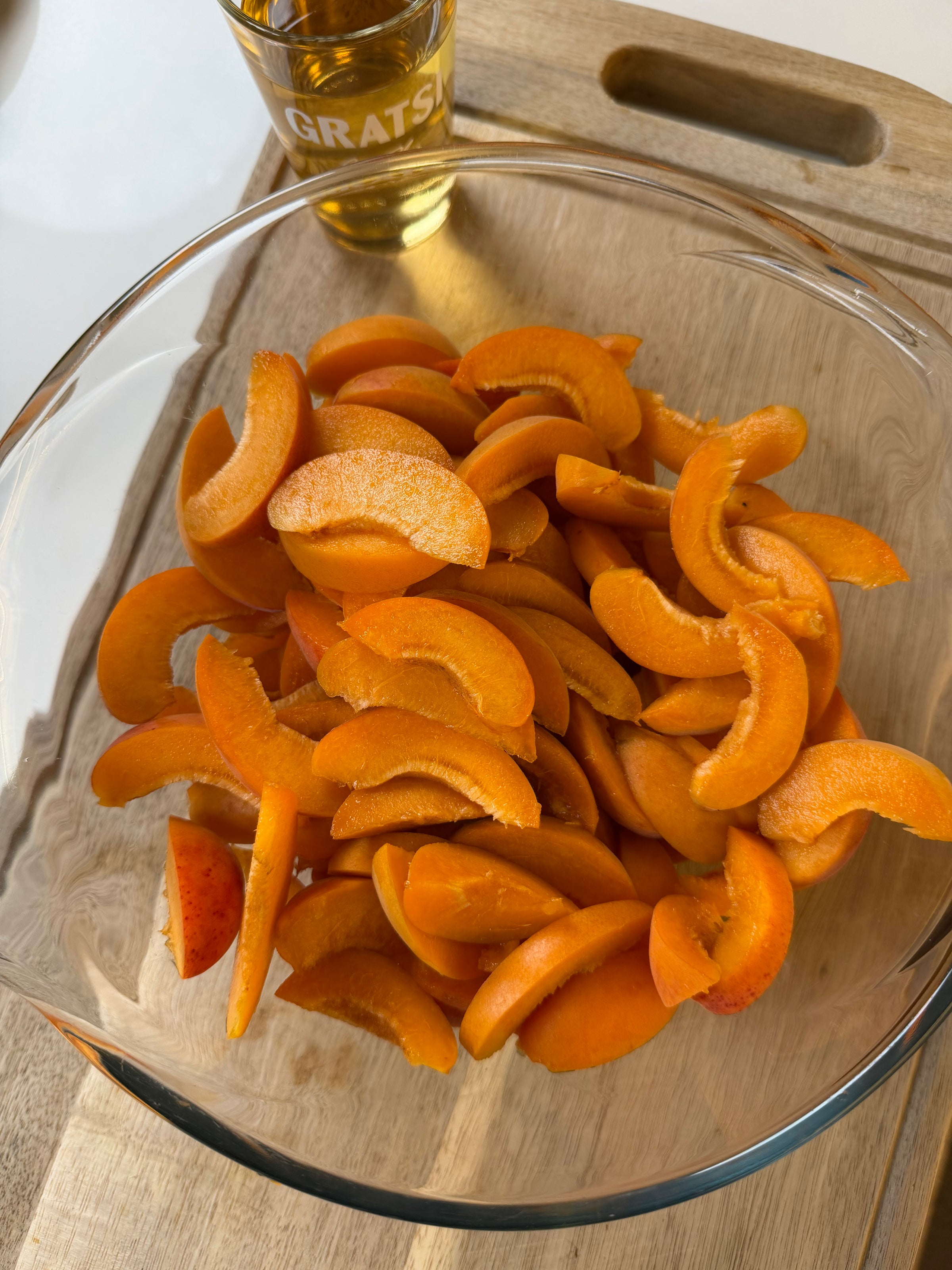 A glass bowl filled with sliced apricots sits on a wooden surface. In the background, there is a glass filled with a yellow liquid.