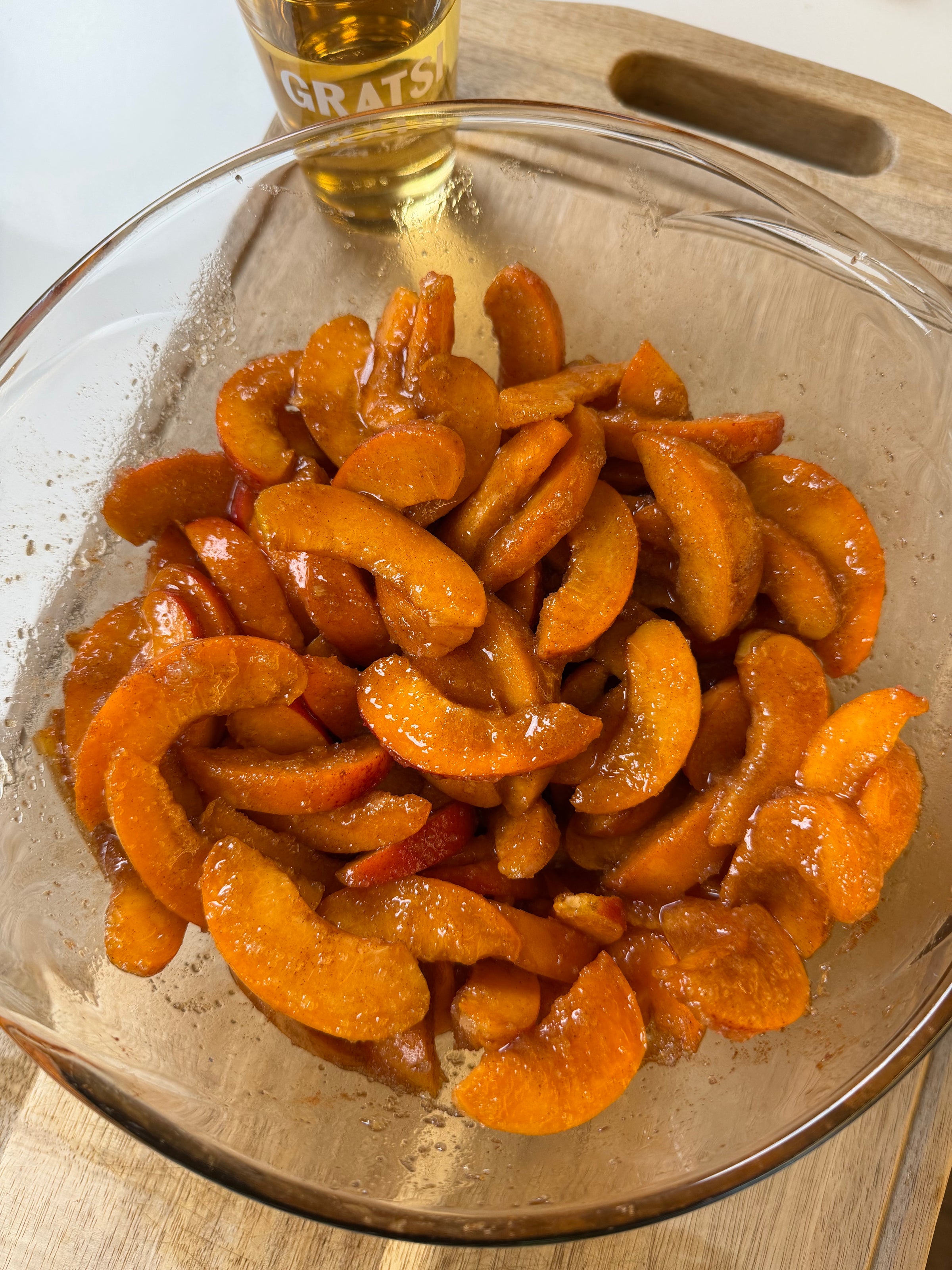Sliced apricots coated in sugar or syrup in a glass bowl on a wooden cutting board, with a partially visible glass of light-colored drink in the background.