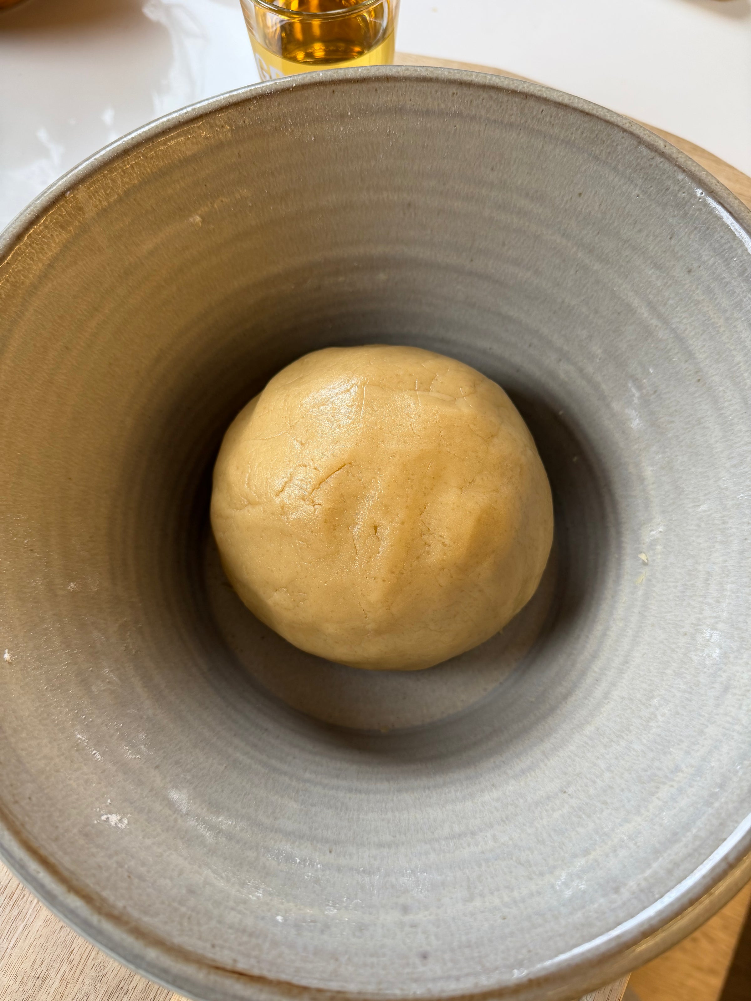 A ball of dough sits in the center of a large, gray ceramic bowl on a wooden surface, ready for baking; a glass with liquid is visible in the background.