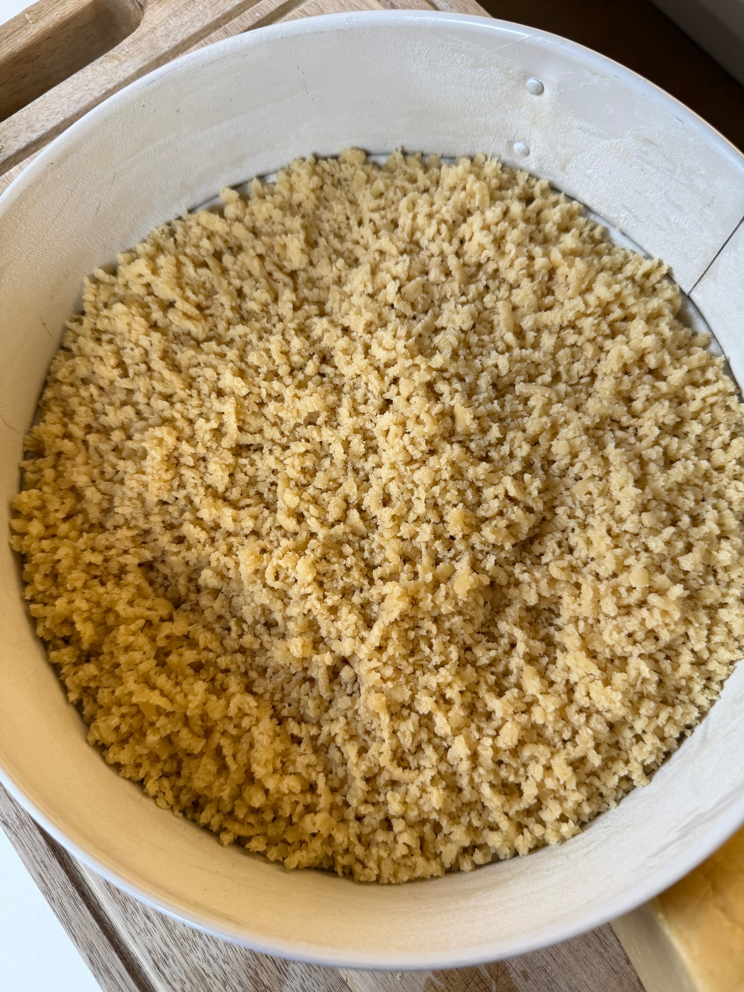 A close-up of a round baking pan filled with an even layer of uncooked crumbly dough, likely for a crust or crumble topping, placed on a wooden surface.