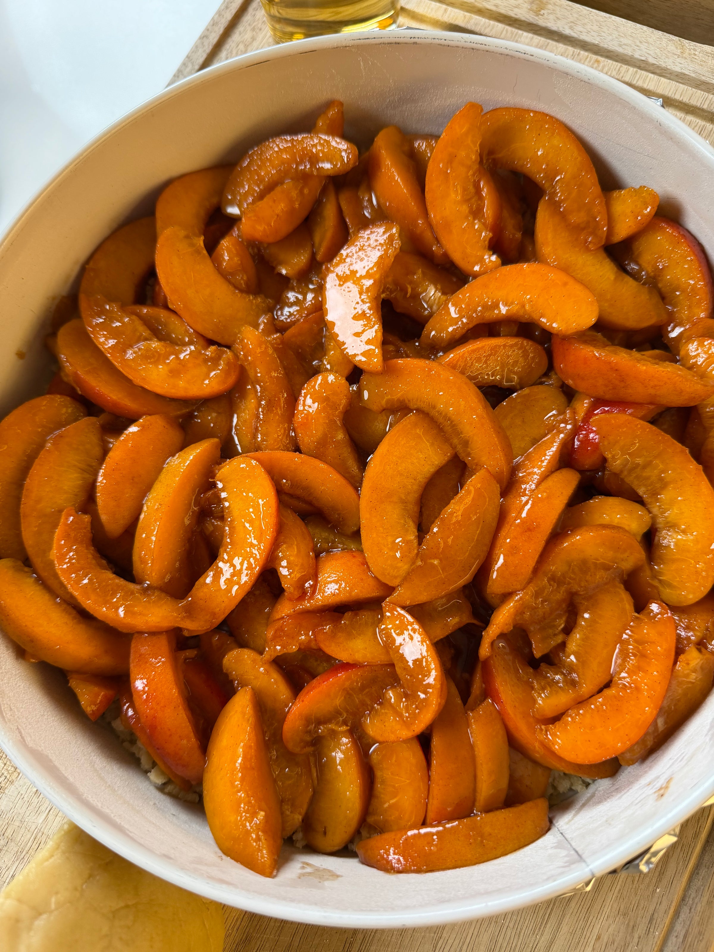 Sliced peaches coated in a glossy syrup are arranged in a white bowl on a wooden surface, ready to be used for baking or eating.