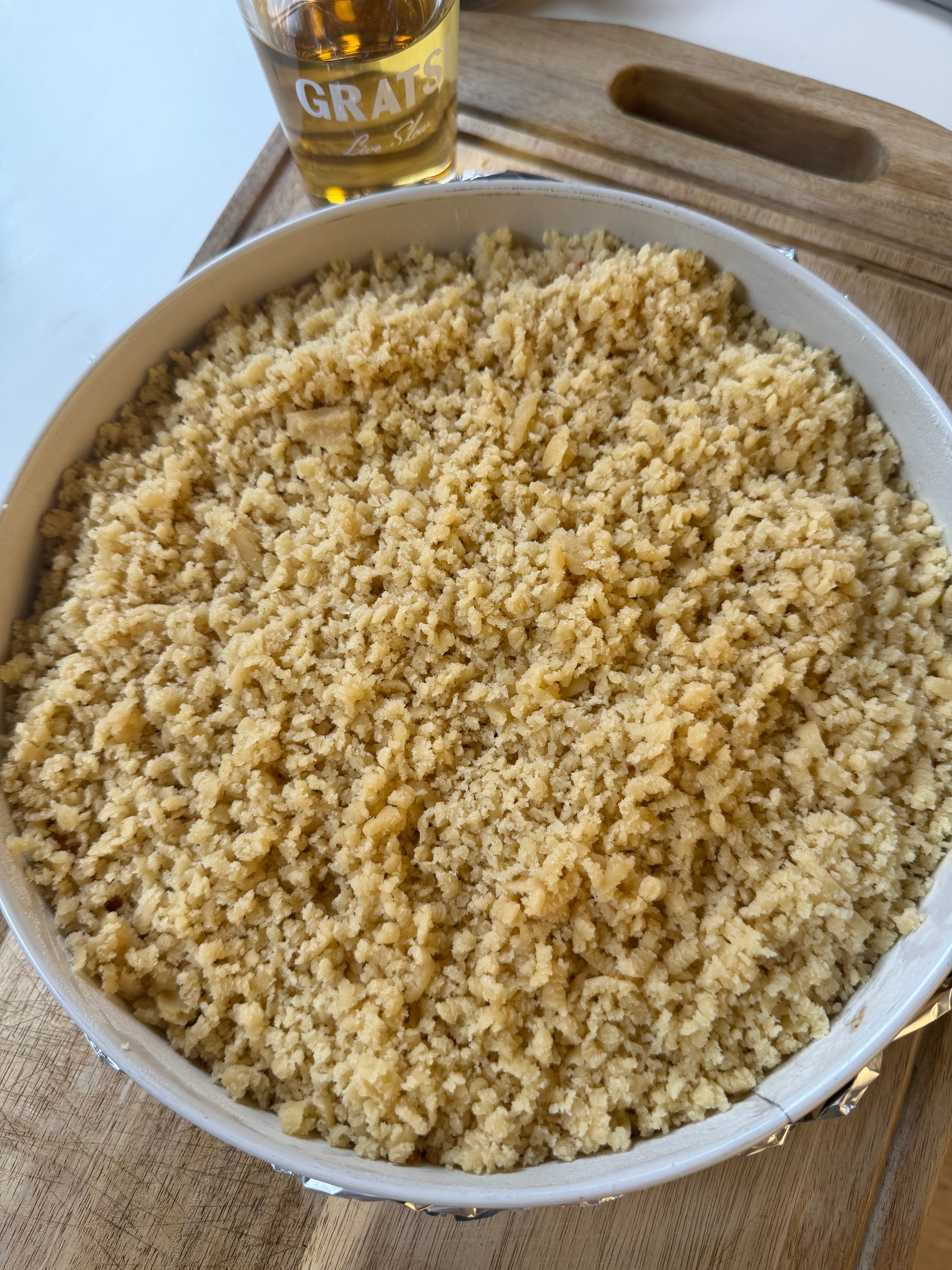 A round baking dish filled with an uncooked crumb topping sits on a wooden cutting board, with a glass of amber liquid in the background.