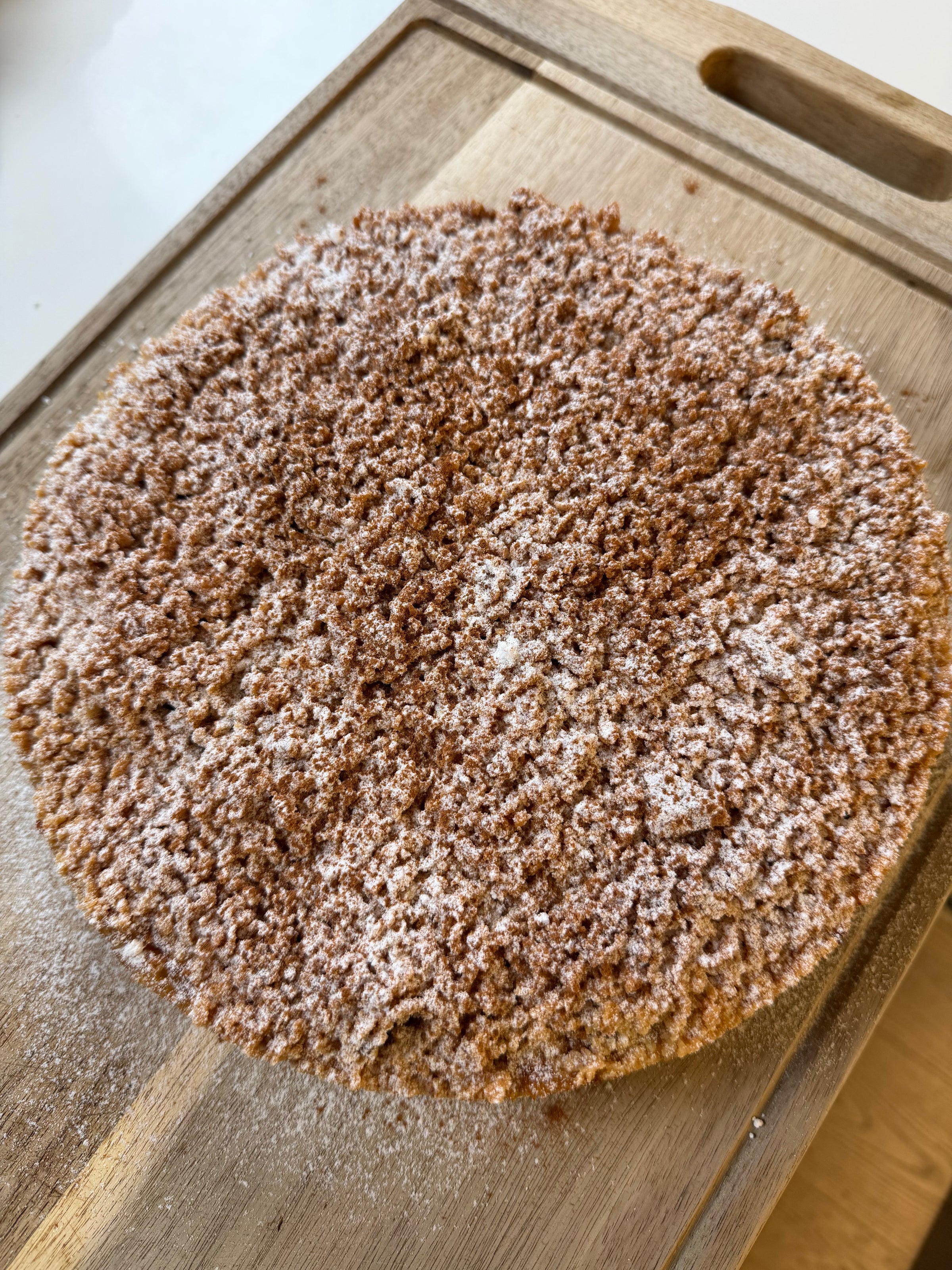 A round cake covered in powdered sugar and crumbled topping sits on a wooden cutting board.