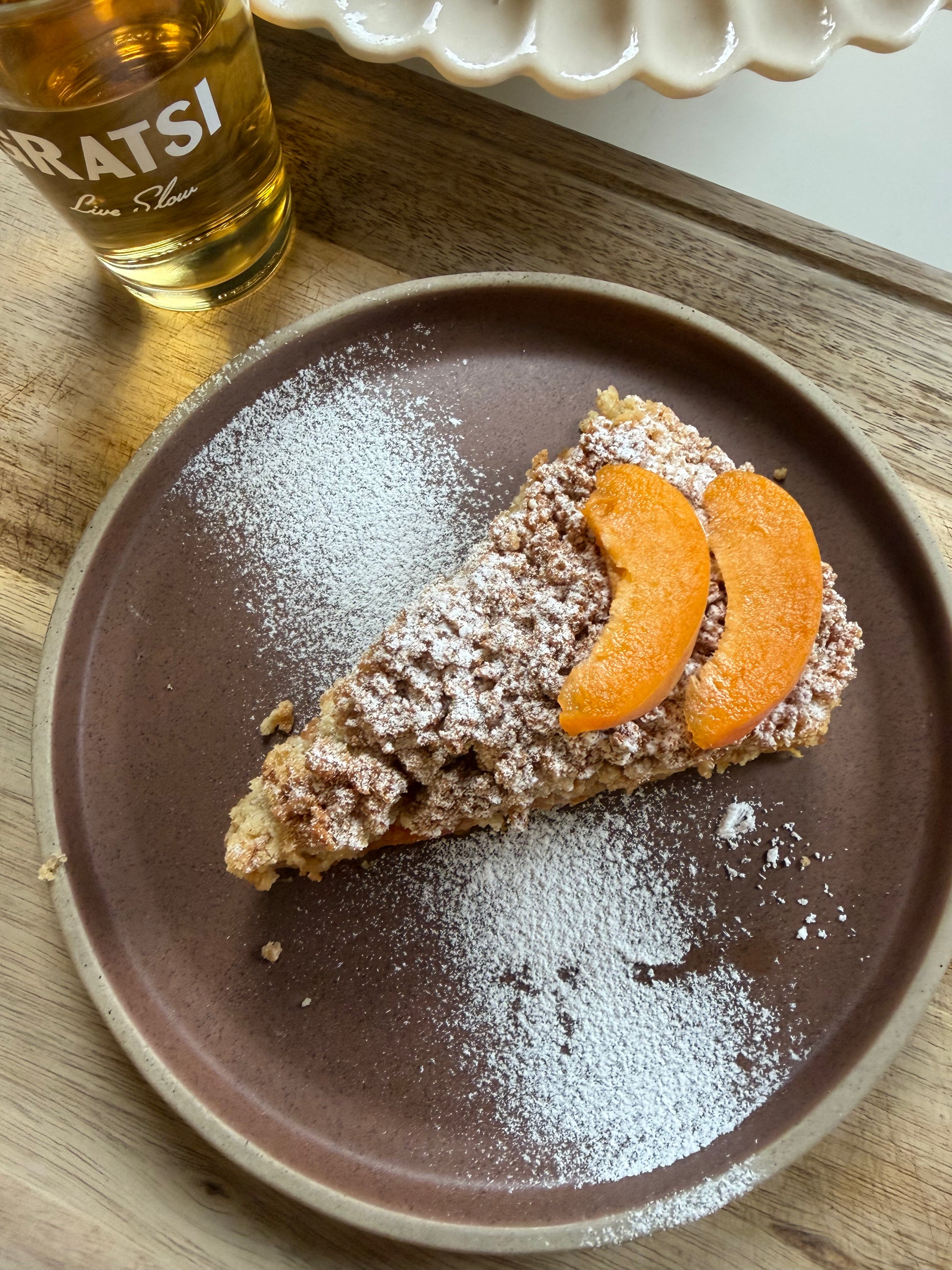 A slice of crumb-topped cake dusted with powdered sugar and garnished with two apricot slices on a brown plate, next to a glass of yellow beverage labeled GRATSI.