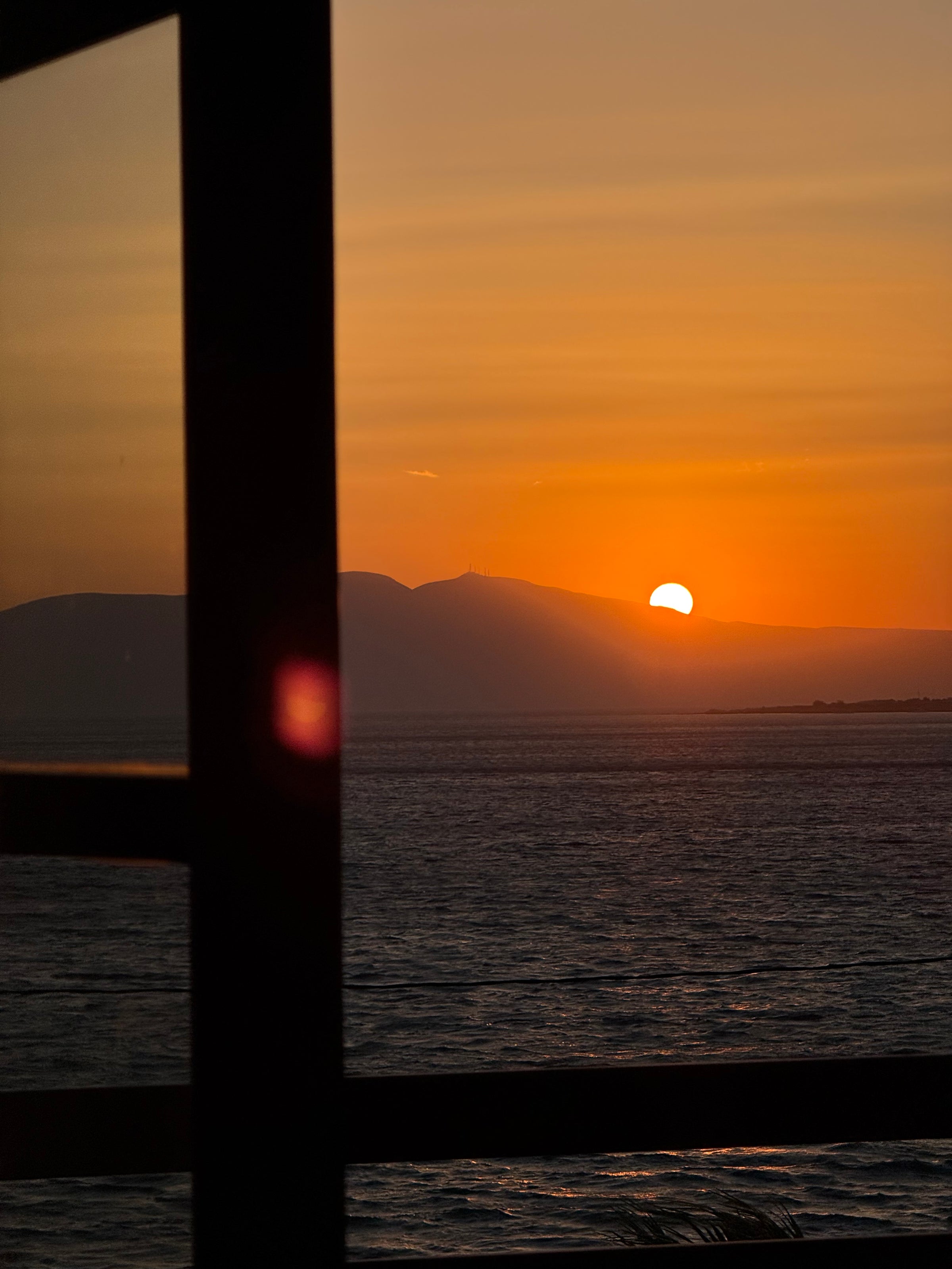 Sunset over calm water with distant mountains in the background, viewed through a window with dark silhouetted frames. The orange sky reflects on the surface of the water.