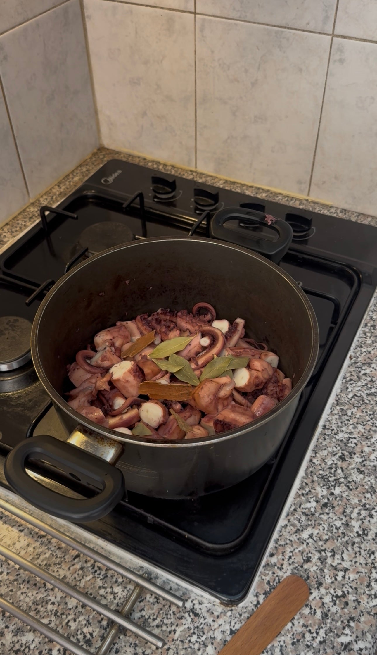 A pot on a stovetop contains chopped squid, garlic cloves, and bay leaves cooking together. The kitchen counter and tiled wall are visible in the background.