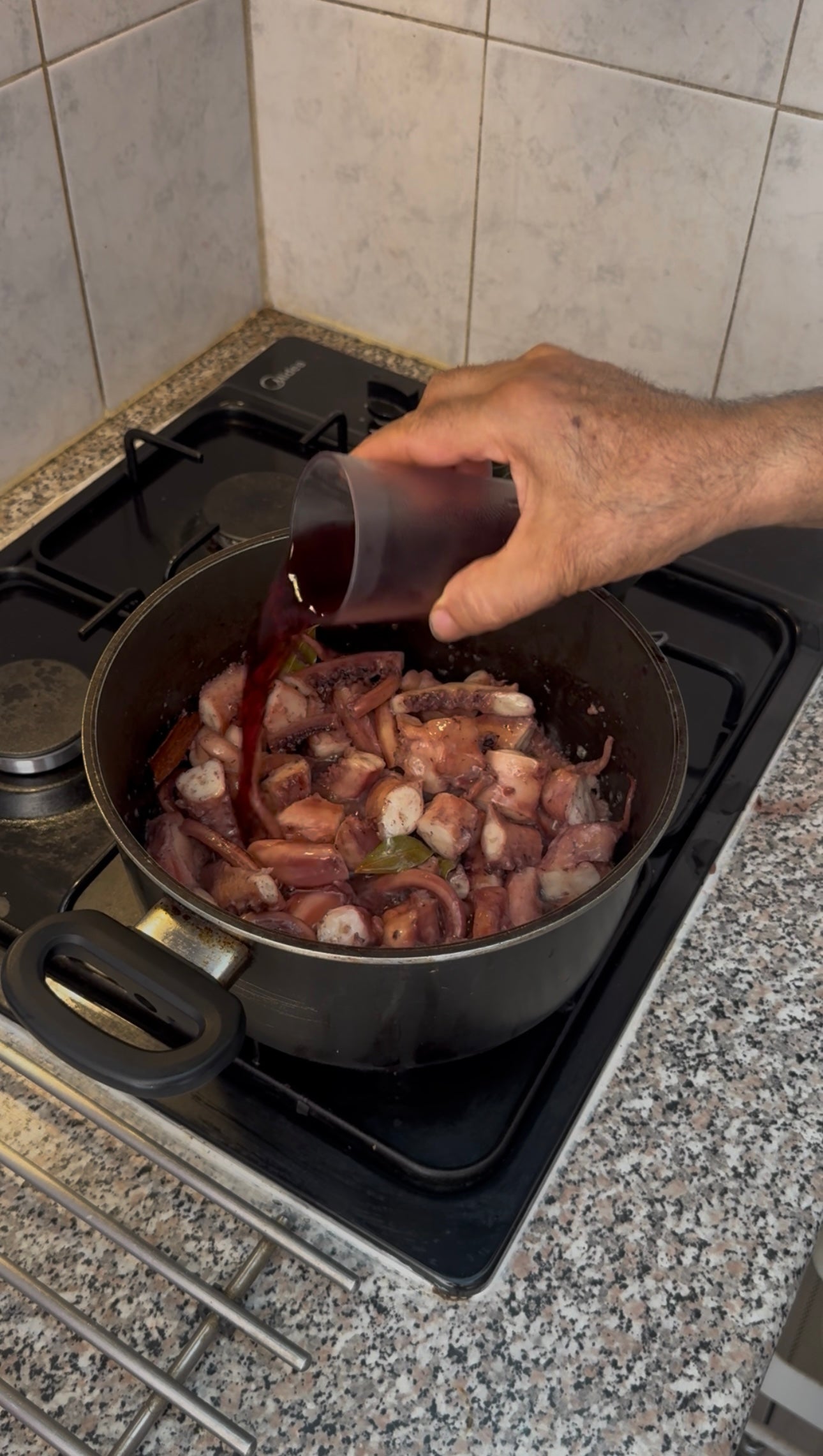 A hand pours red wine from a small cup into a pan of octopus cooking on a stove, surrounded by kitchen tiles and a granite countertop.