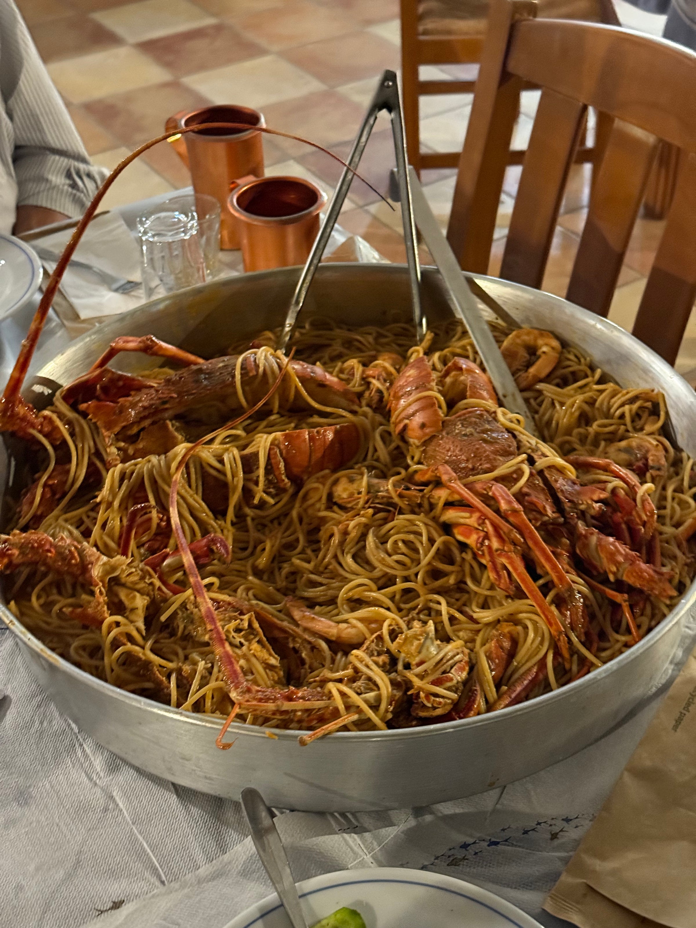 A large metal pan filled with spaghetti and several whole lobsters sits on a table set for a meal, with tongs resting on top.