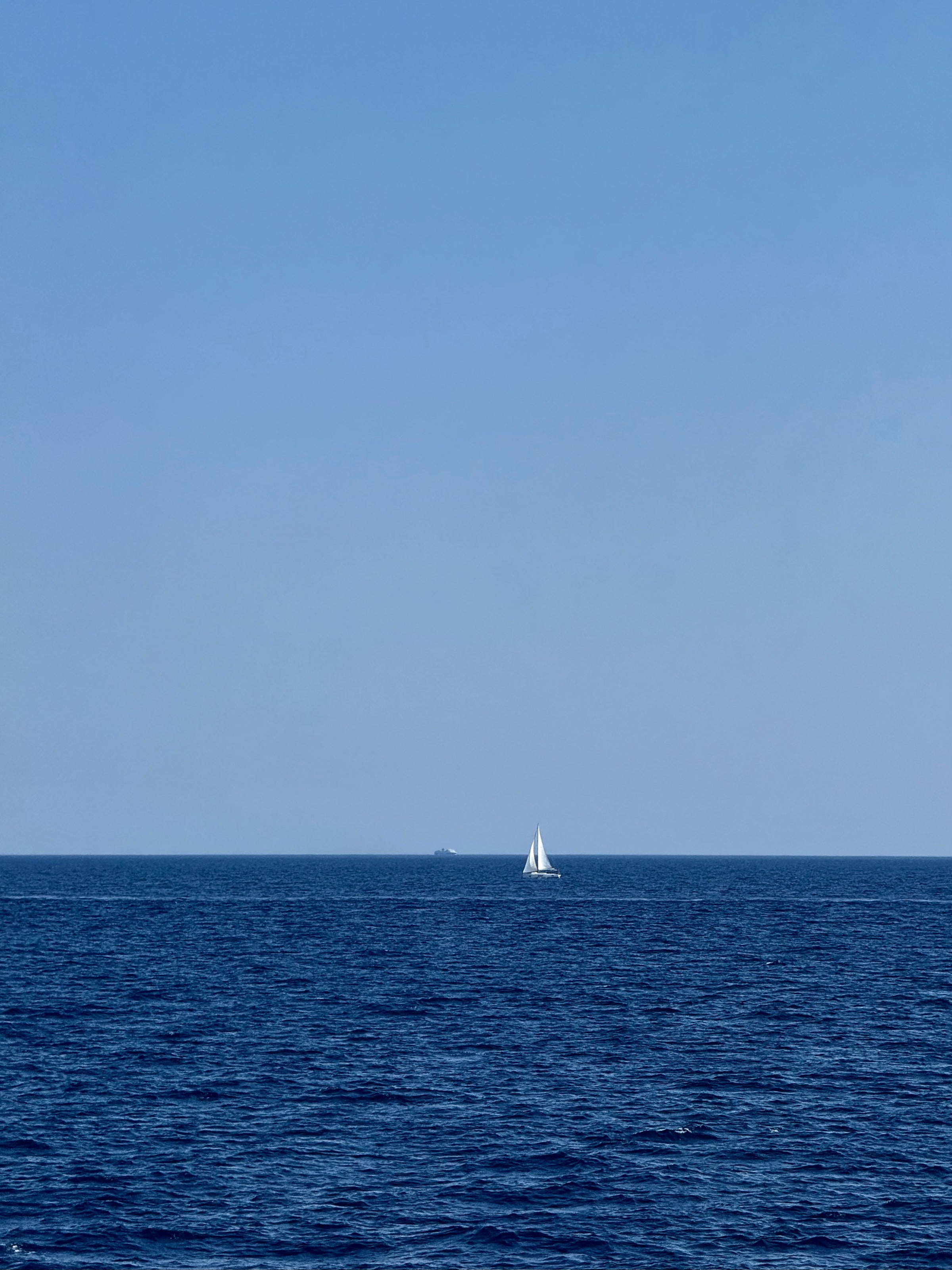 A small white sailboat floats on calm, deep blue water under a clear blue sky, with the horizon visible in the distance.