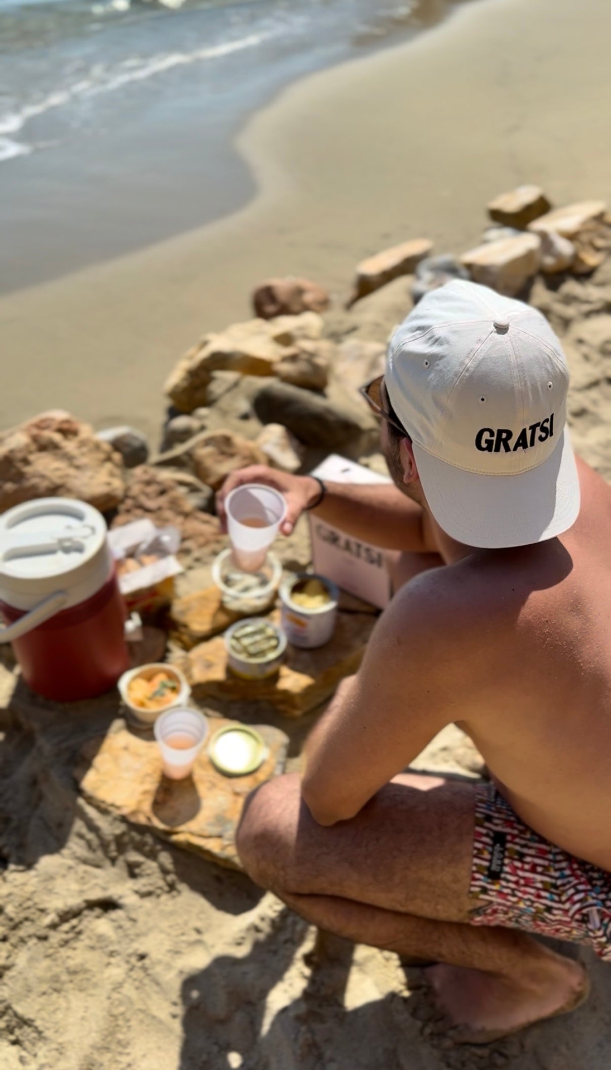 A person wearing a white “GRATSI” hat sits on rocks at the beach, holding a cup next to snacks, dips, and a drink cooler. The ocean is visible in the background.