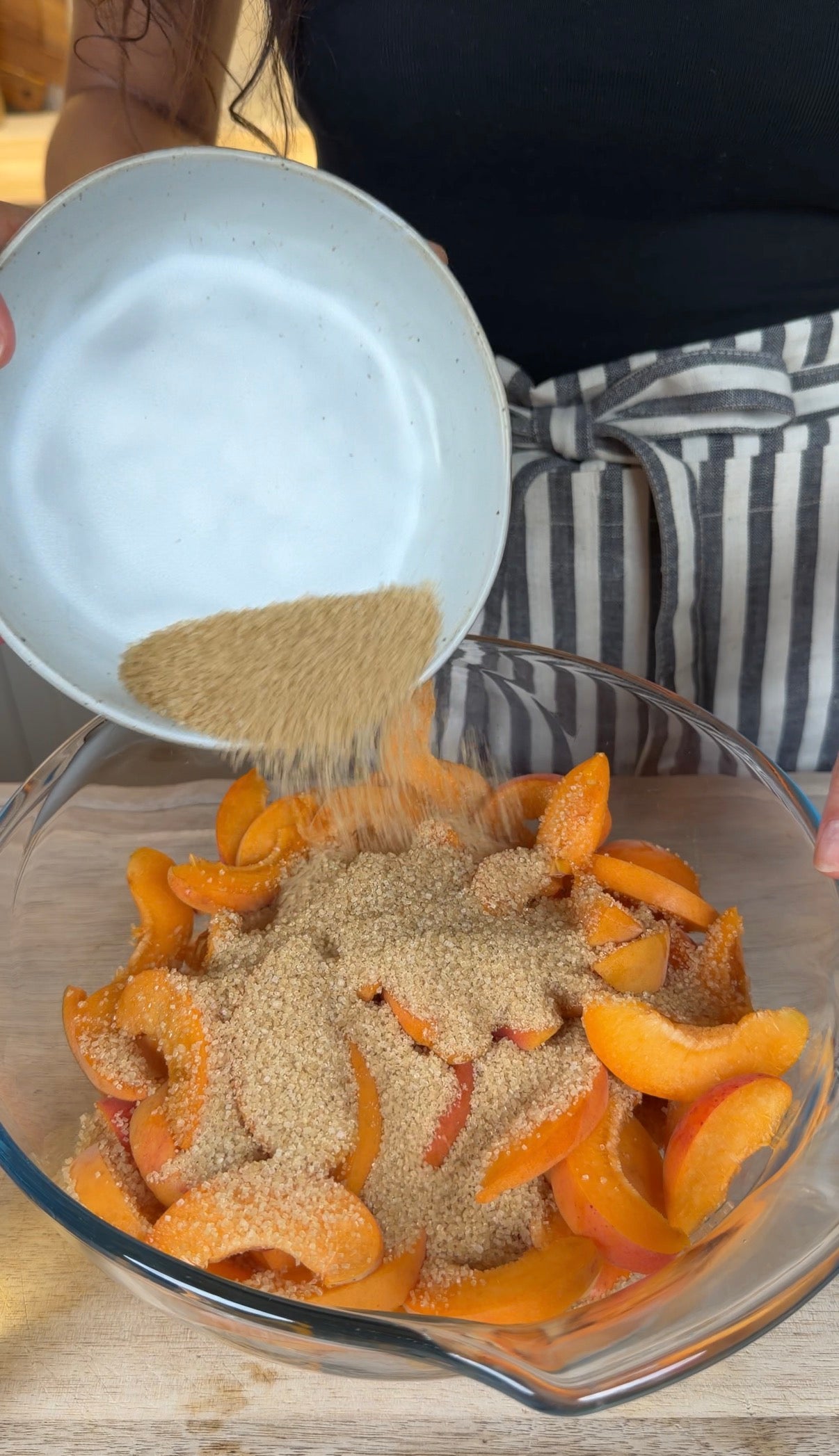 A person pours brown sugar from a white bowl over sliced peaches in a glass mixing bowl. They are wearing a striped apron.