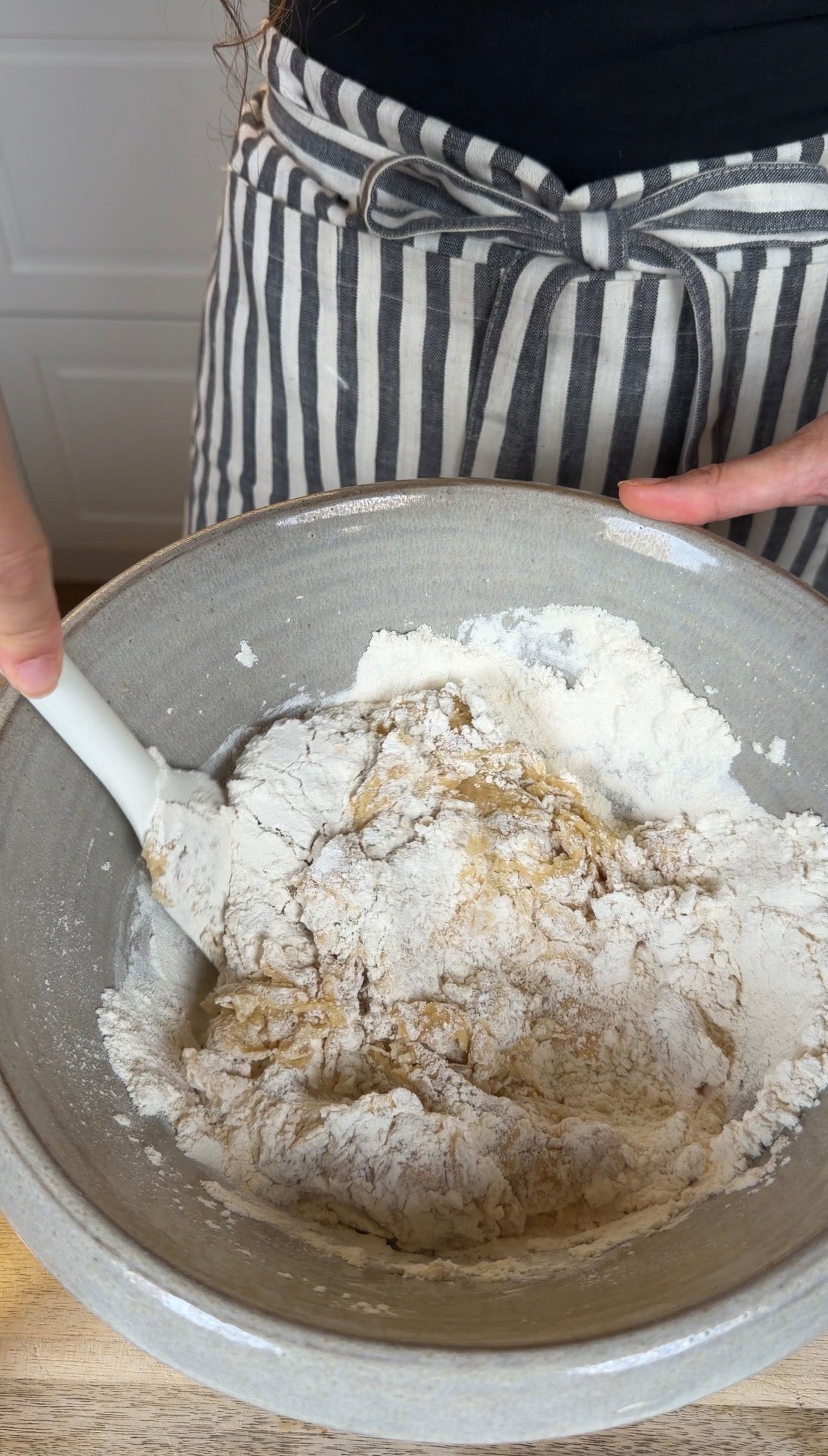 Person in a striped apron mixing flour and other ingredients in a large gray bowl with a white spatula, preparing dough.