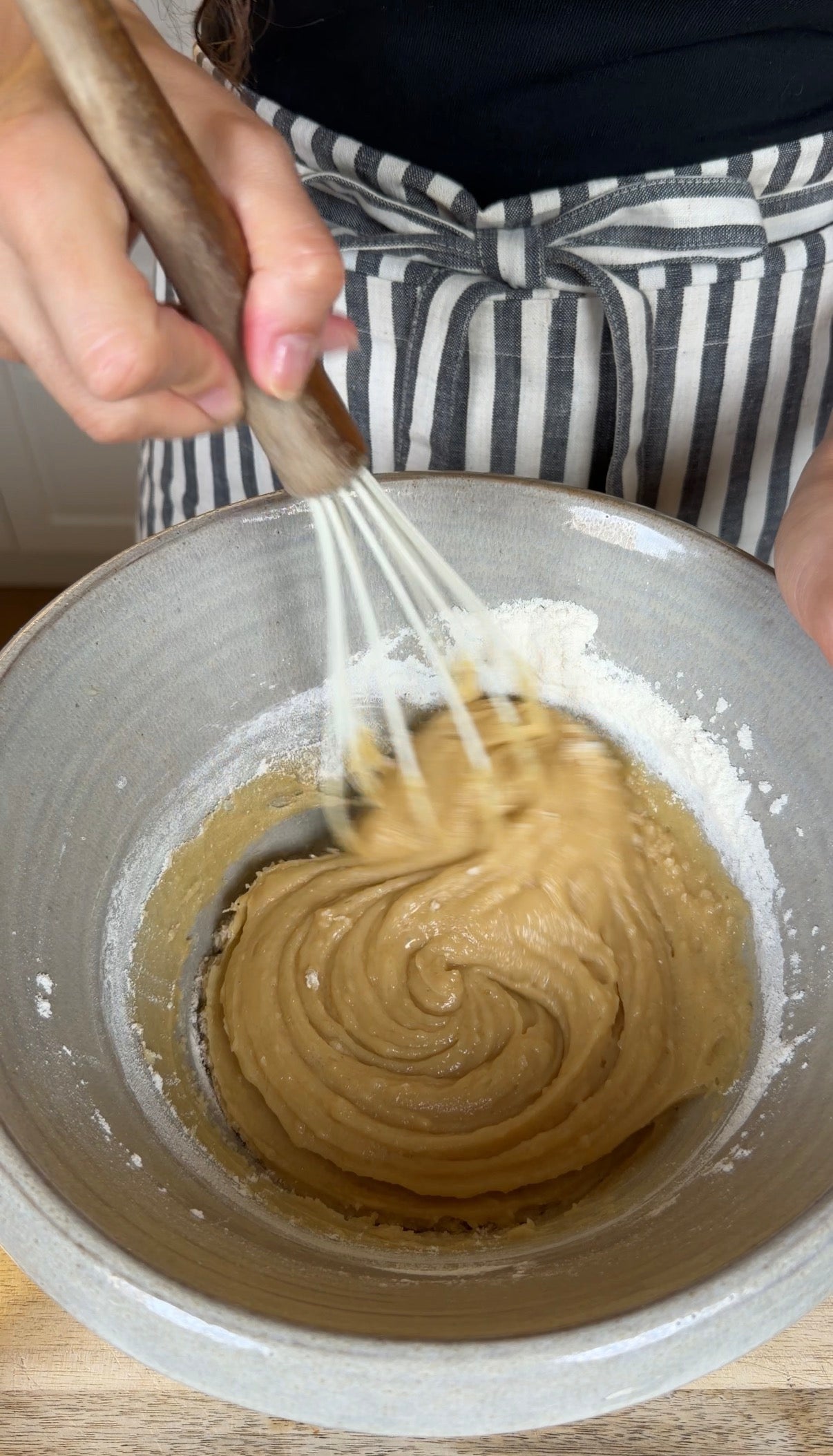 A person wearing a striped apron mixes thick, light brown batter in a gray bowl with a whisk. Flour is visible along the sides of the bowl.
