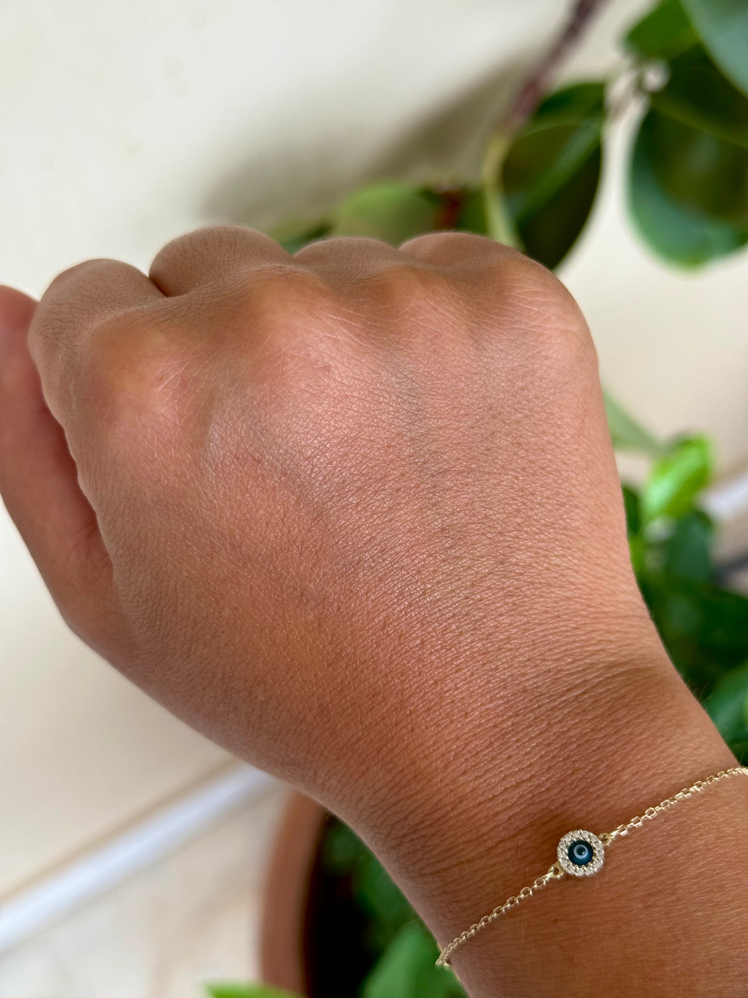 A hand with a gold chain bracelet featuring a small, round evil eye charm is shown against a background of green leaves and a beige wall.