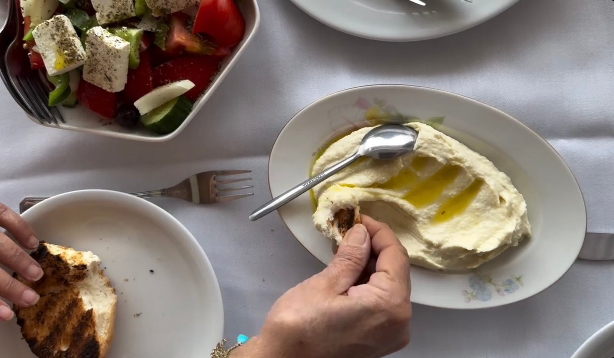A person dips grilled bread into a plate of creamy spread drizzled with olive oil, next to a Greek salad with tomatoes, cucumbers, olives, and feta on a white tablecloth.