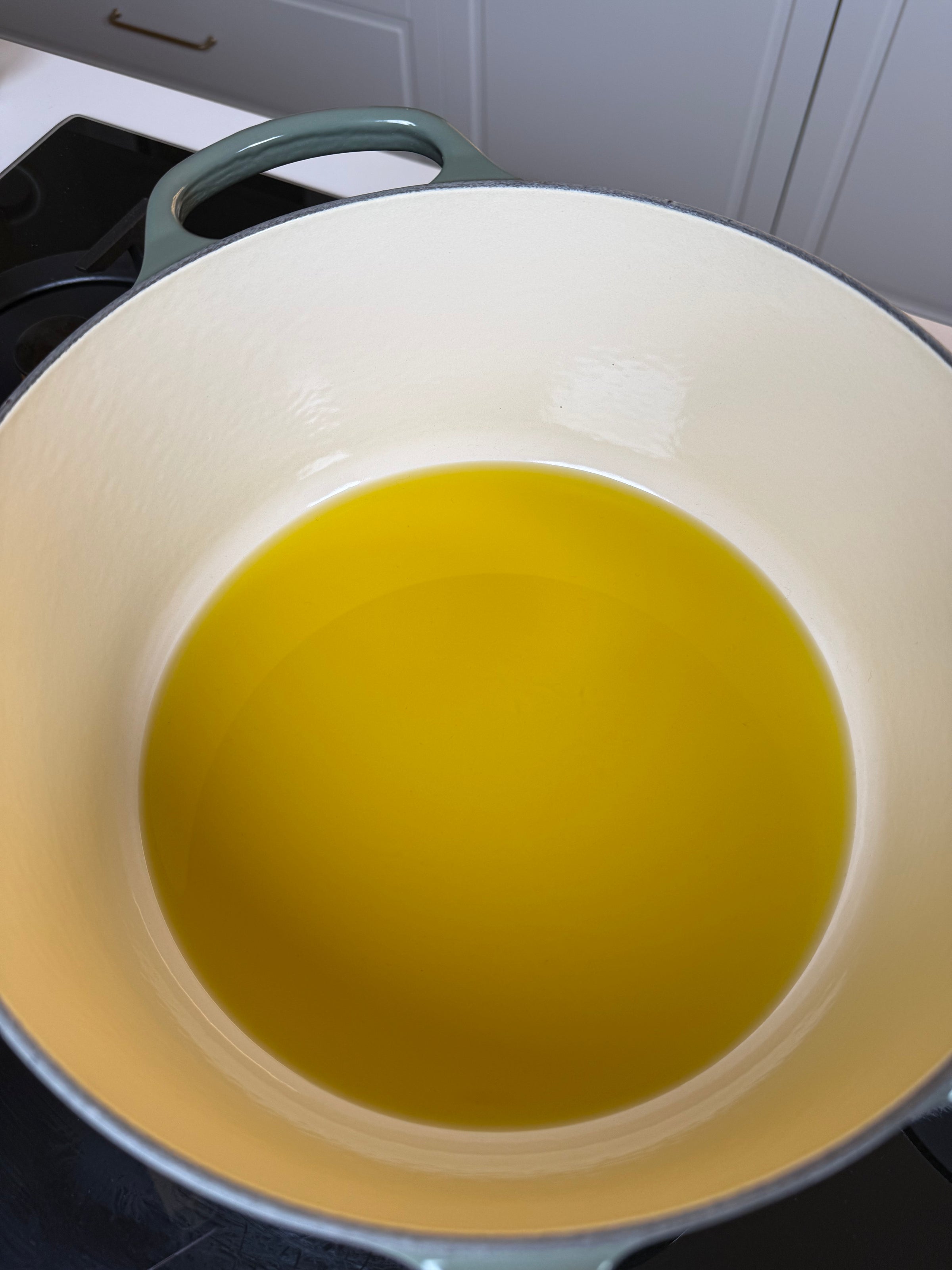 A green Dutch oven sits on a stovetop, filled with a layer of yellow cooking oil. White cabinets and a dark countertop are visible in the background.