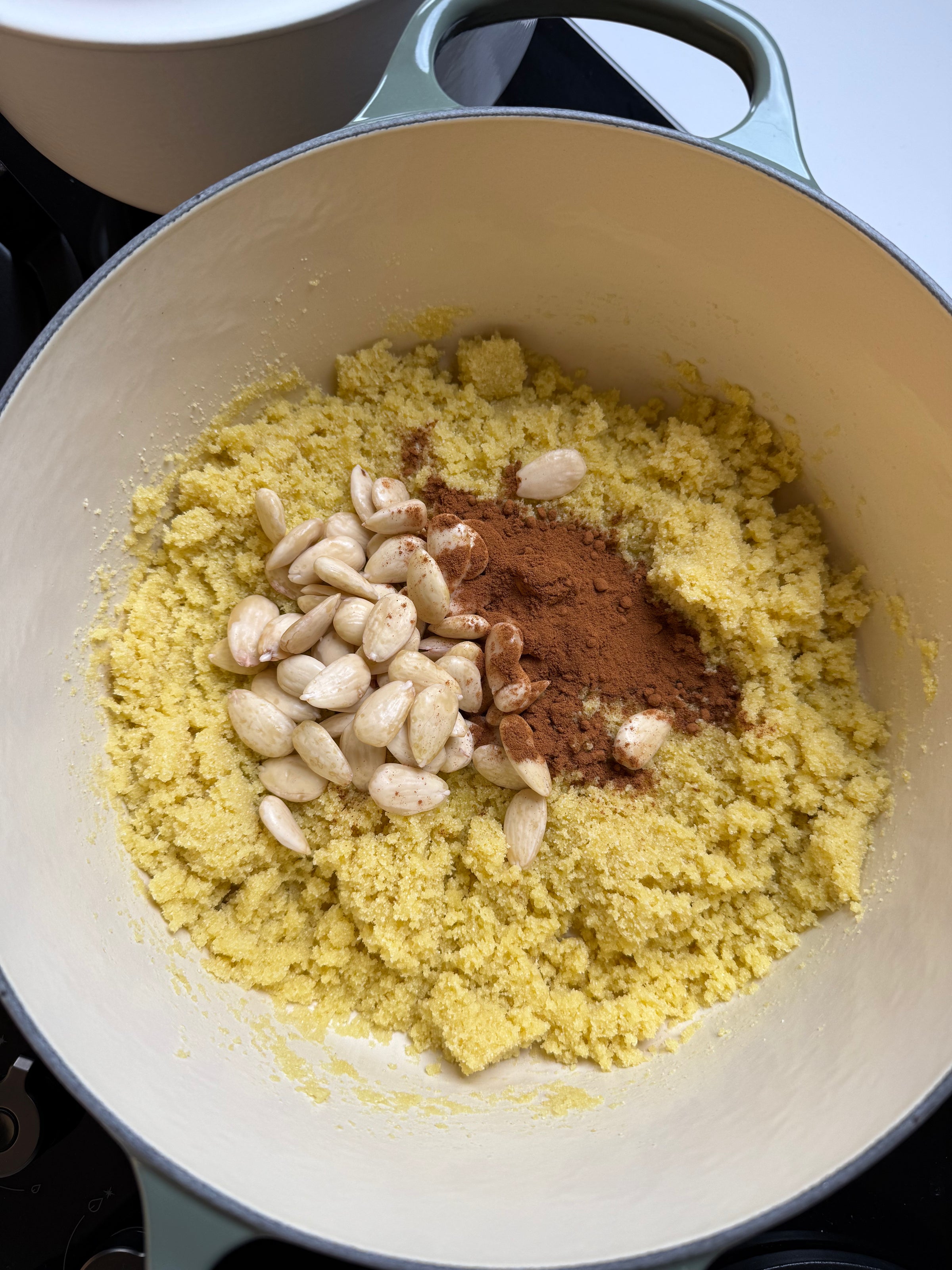 A pot filled with a yellow, crumbly mixture, topped with blanched almonds and a pile of ground cinnamon, ready to be mixed.