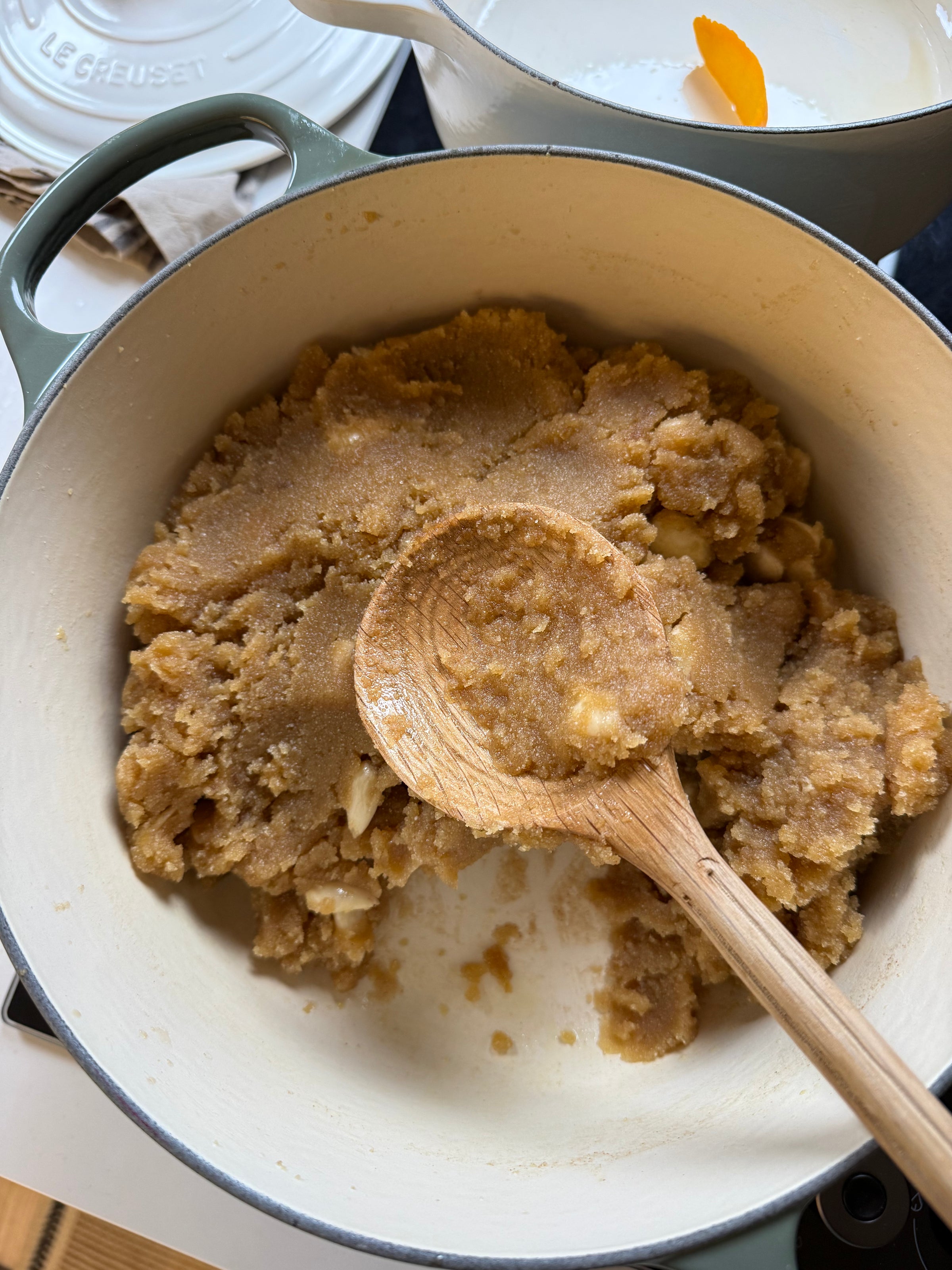 A wooden spoon rests in a pot filled with a crumbly, brown, and grainy mixture, possibly a dessert or dough, inside a light-colored Dutch oven on a stovetop.