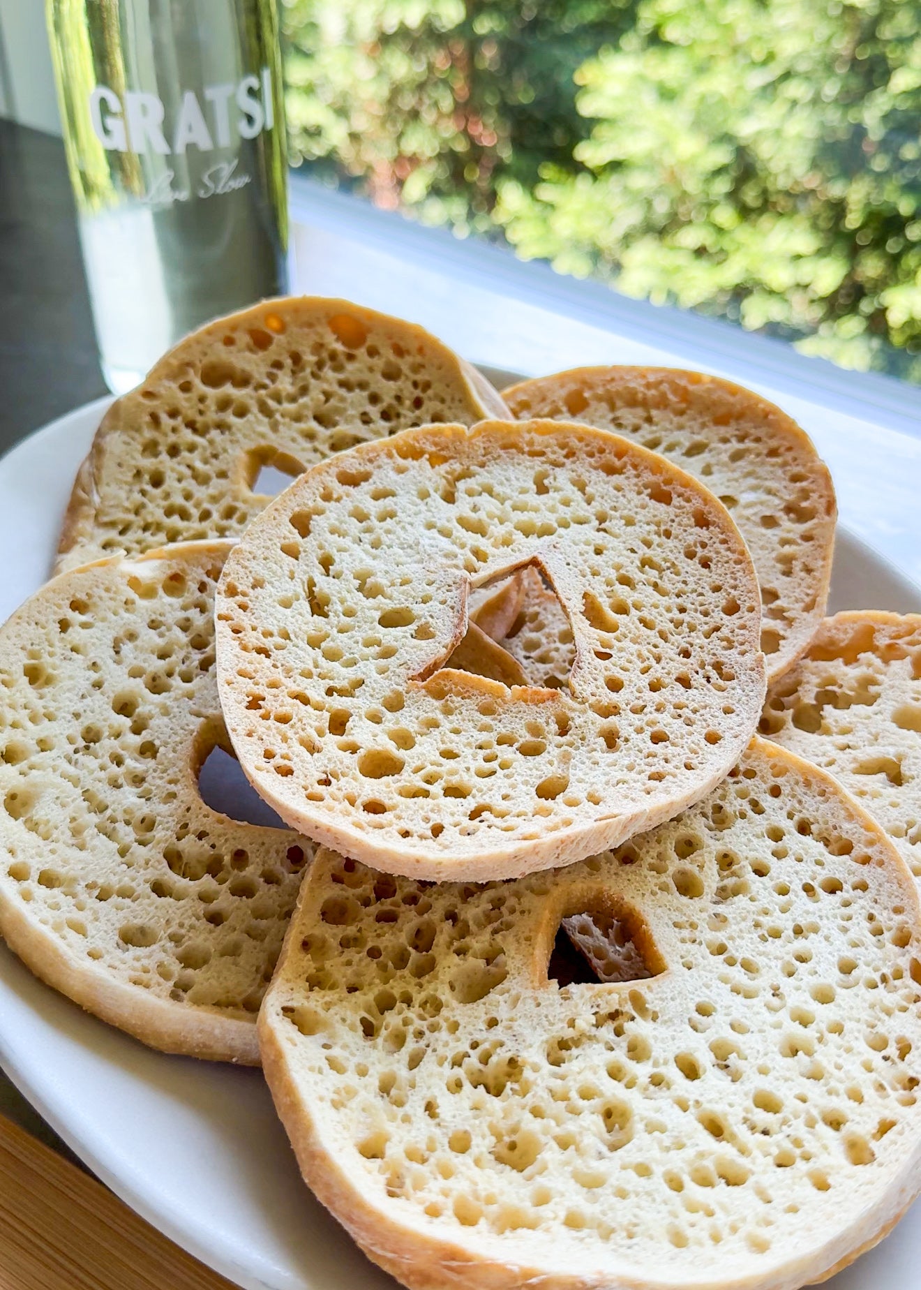 A plate of sliced crumpets with holes in the middle, sitting on a white surface near a window. A bottle labeled GRATSI is in the background, and green foliage is visible outside.