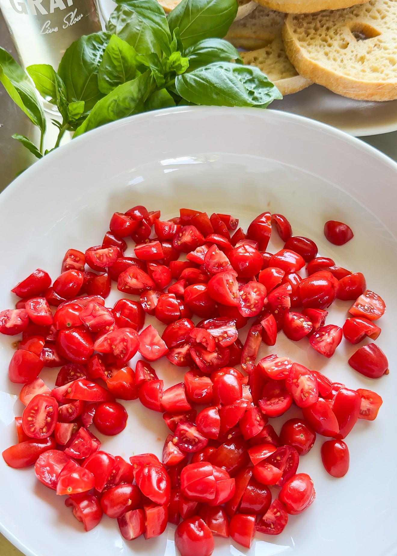 Chopped cherry tomatoes on a white plate, with fresh basil leaves and slices of bread in the background.