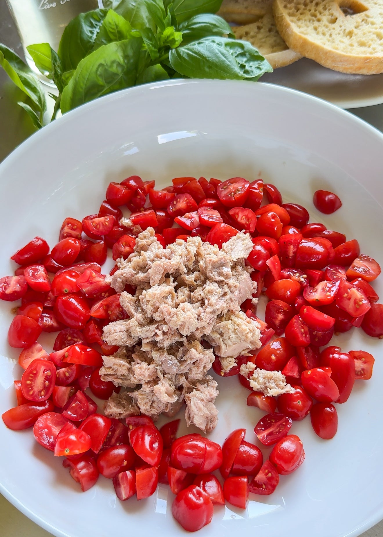 A white bowl with halved cherry tomatoes surrounding a pile of tuna. Fresh basil leaves and slices of bread are in the background.