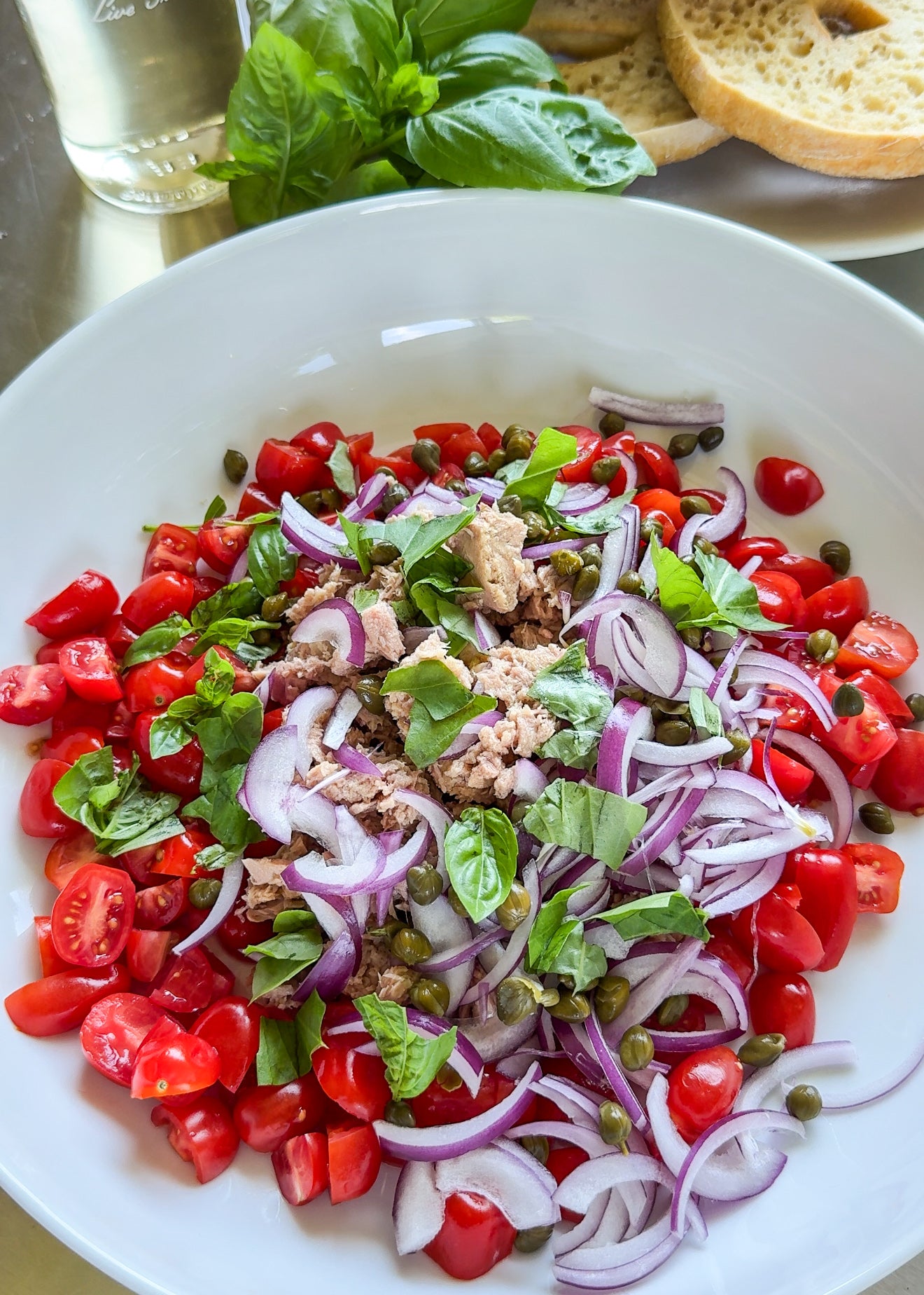 A white bowl filled with chopped cherry tomatoes, sliced red onions, tuna, capers, and fresh basil leaves. Slices of bread, a glass of water, and more basil are in the background.