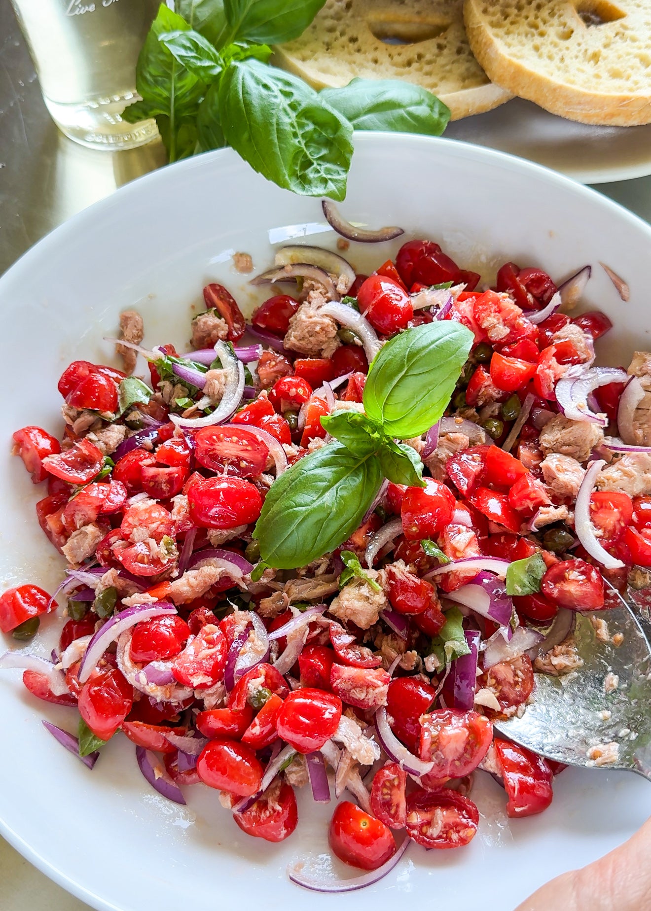A white bowl holds a salad of cherry tomatoes, red onions, tuna, capers, and basil, with bread slices and a basil sprig in the background. A spoon rests in the bowl, and a person’s hand is partially visible.