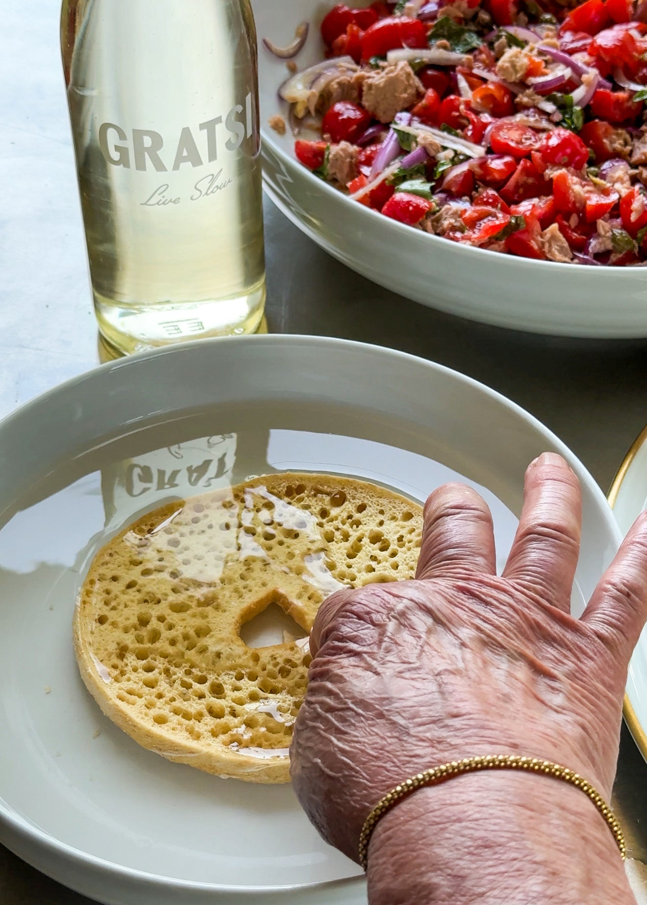 A hand reaches for a spongy round bread dipped in honey on a plate, with a salad bowl and a bottle of white wine labeled GRATSI in the background.