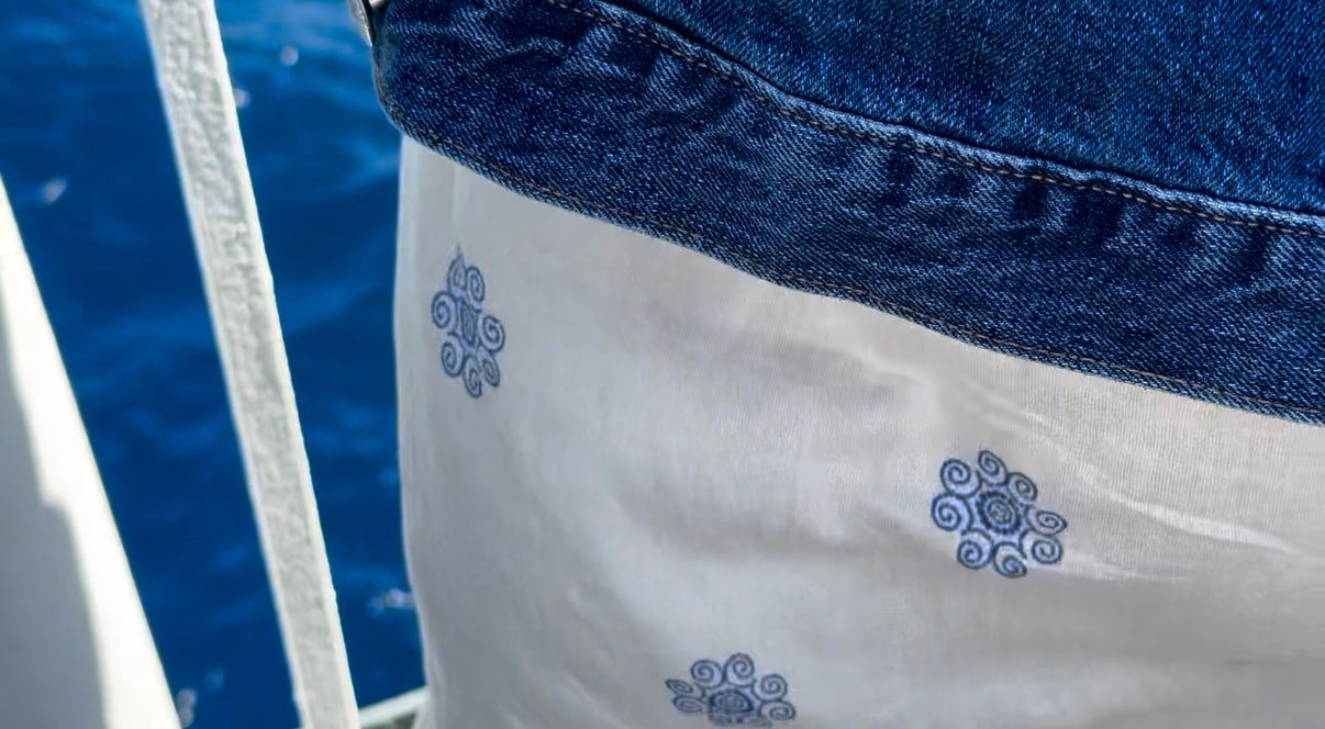 Close-up of a person wearing a blue denim jacket and a white garment with blue swirl patterns, standing near a railing above blue water.