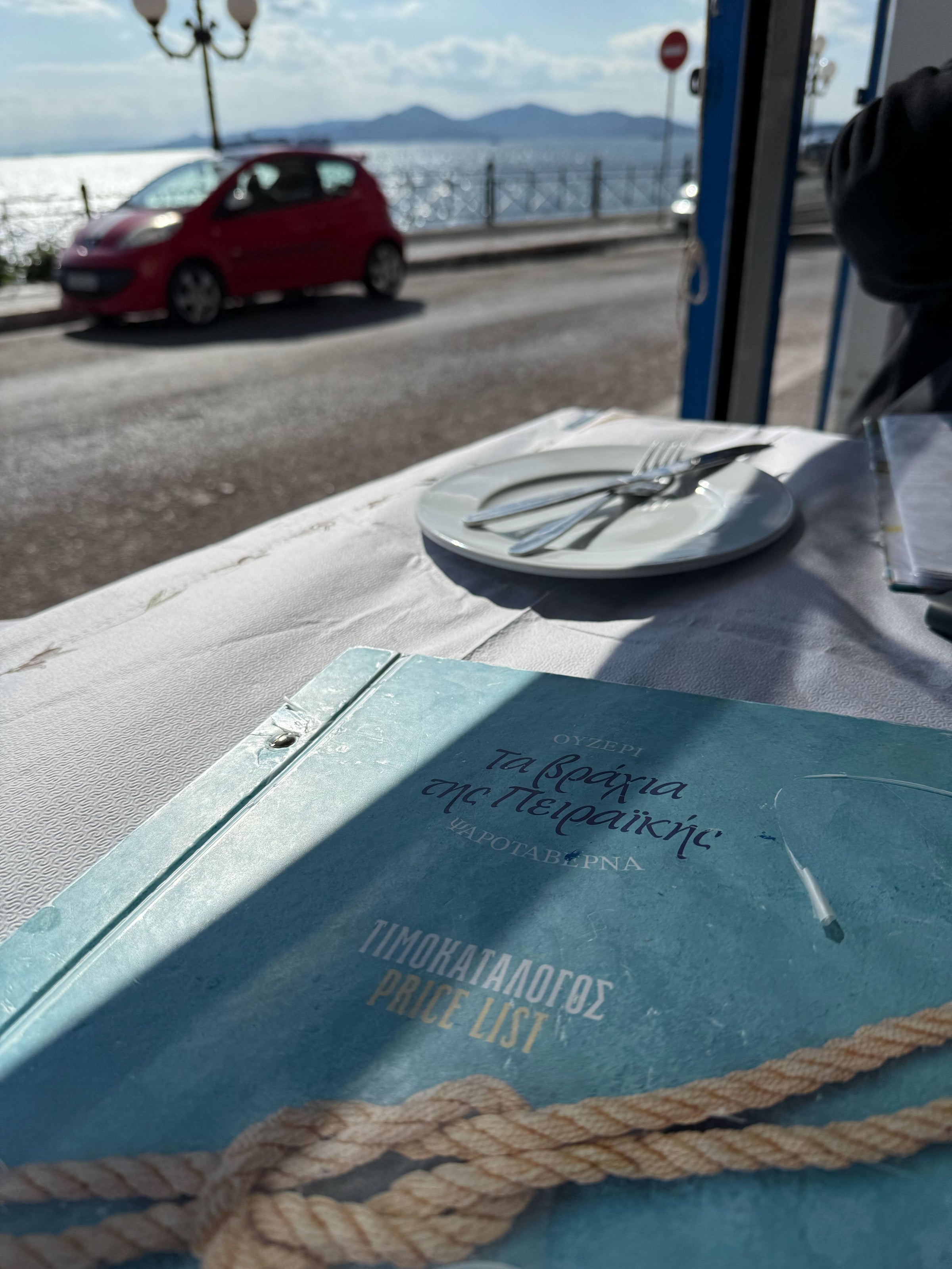 A sunlit seaside view from a restaurant table with a menu in Greek and English in the foreground, a plate with cutlery, and a red car parked by the waterfront road with the sea and mountains in the background.