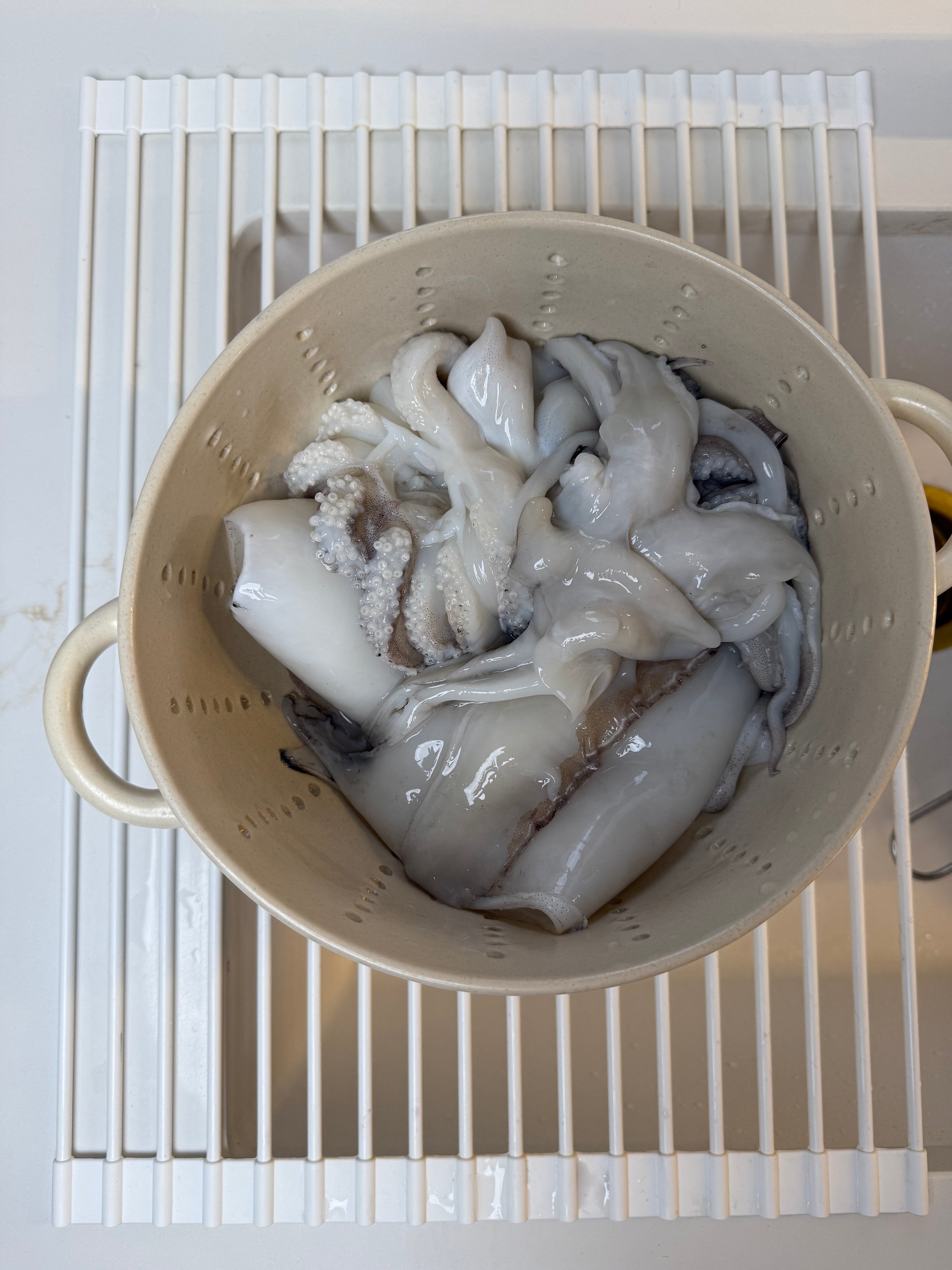 A colander filled with raw, cleaned squid sits on a white dish rack over a sink. The squid are glossy and pale, with visible tentacles and bodies.