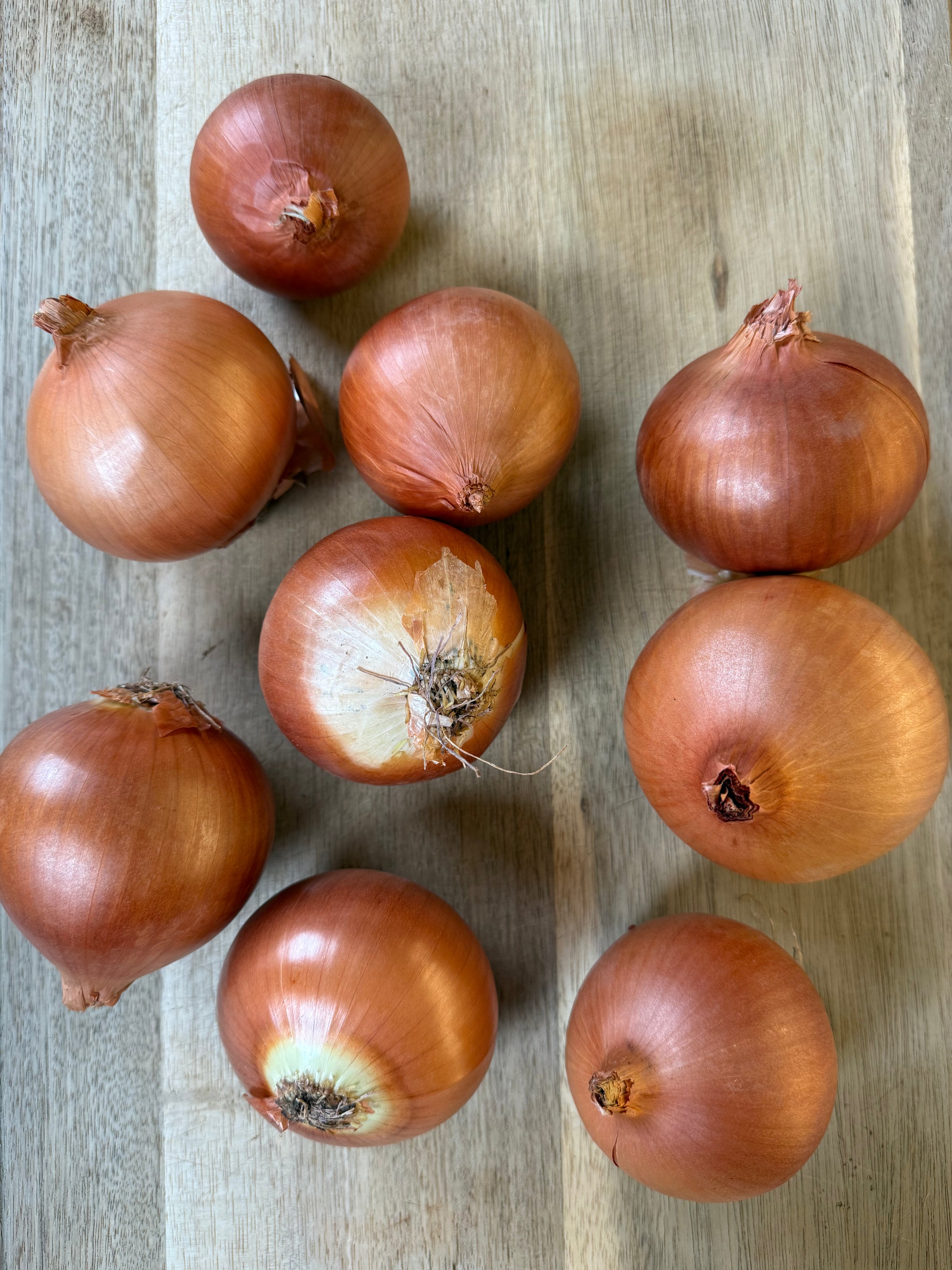 Nine brown onions with papery skins are arranged on a light wooden surface, some with patches of outer skin peeling. The onions vary slightly in size and shape.
