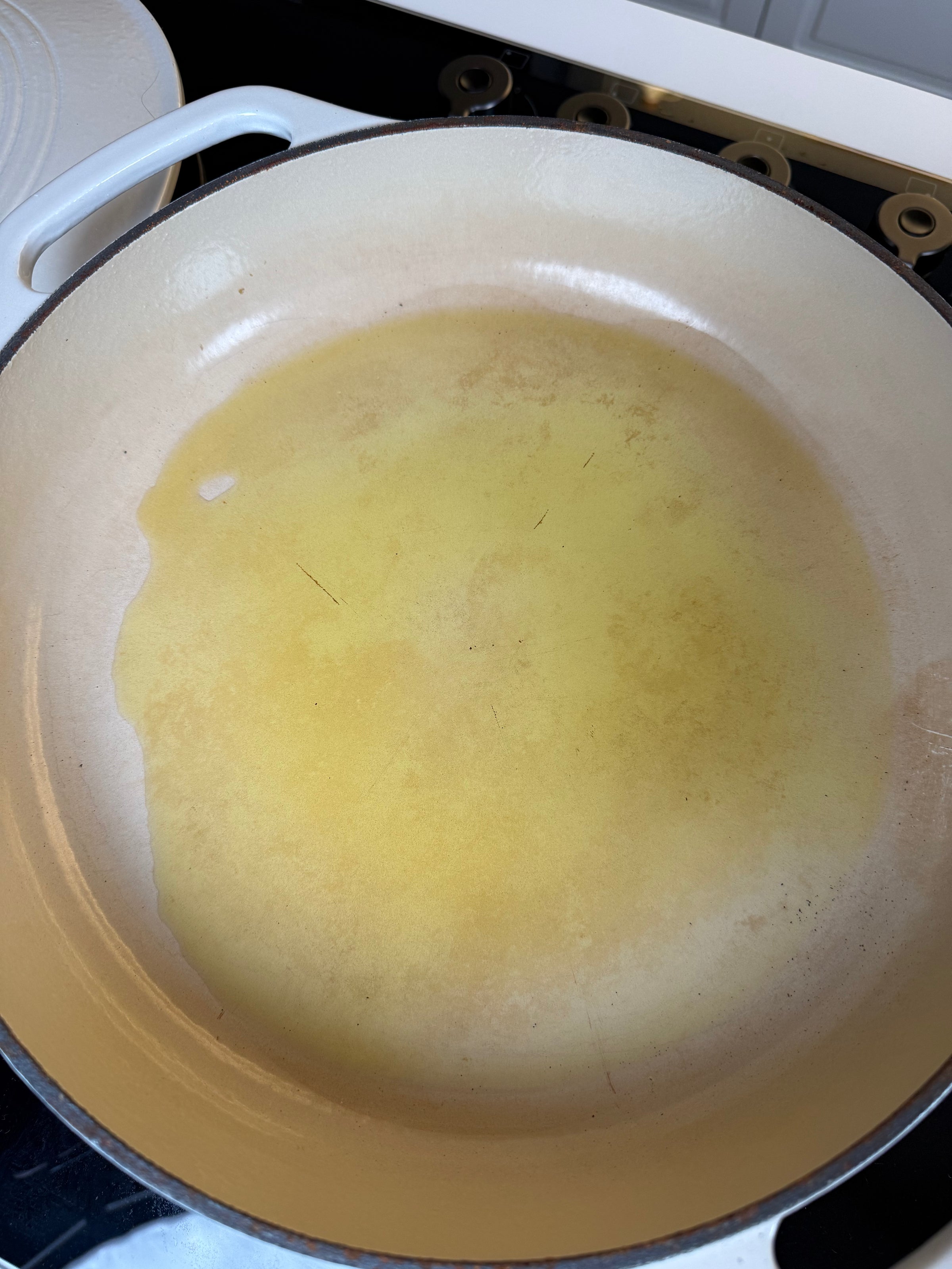 A close-up of a large, beige enameled cast iron pan on a stovetop with a thin layer of oil spread across the bottom, ready for cooking.