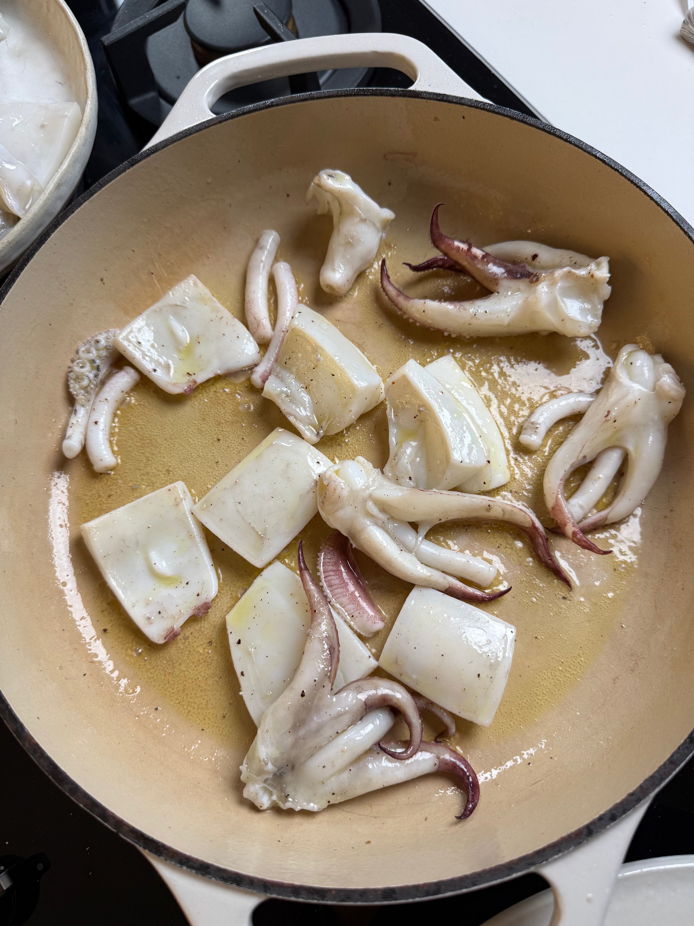 Squid pieces and tentacles are being cooked in a beige pan with some oil. The squid is lightly browned and sizzling on the stovetop.