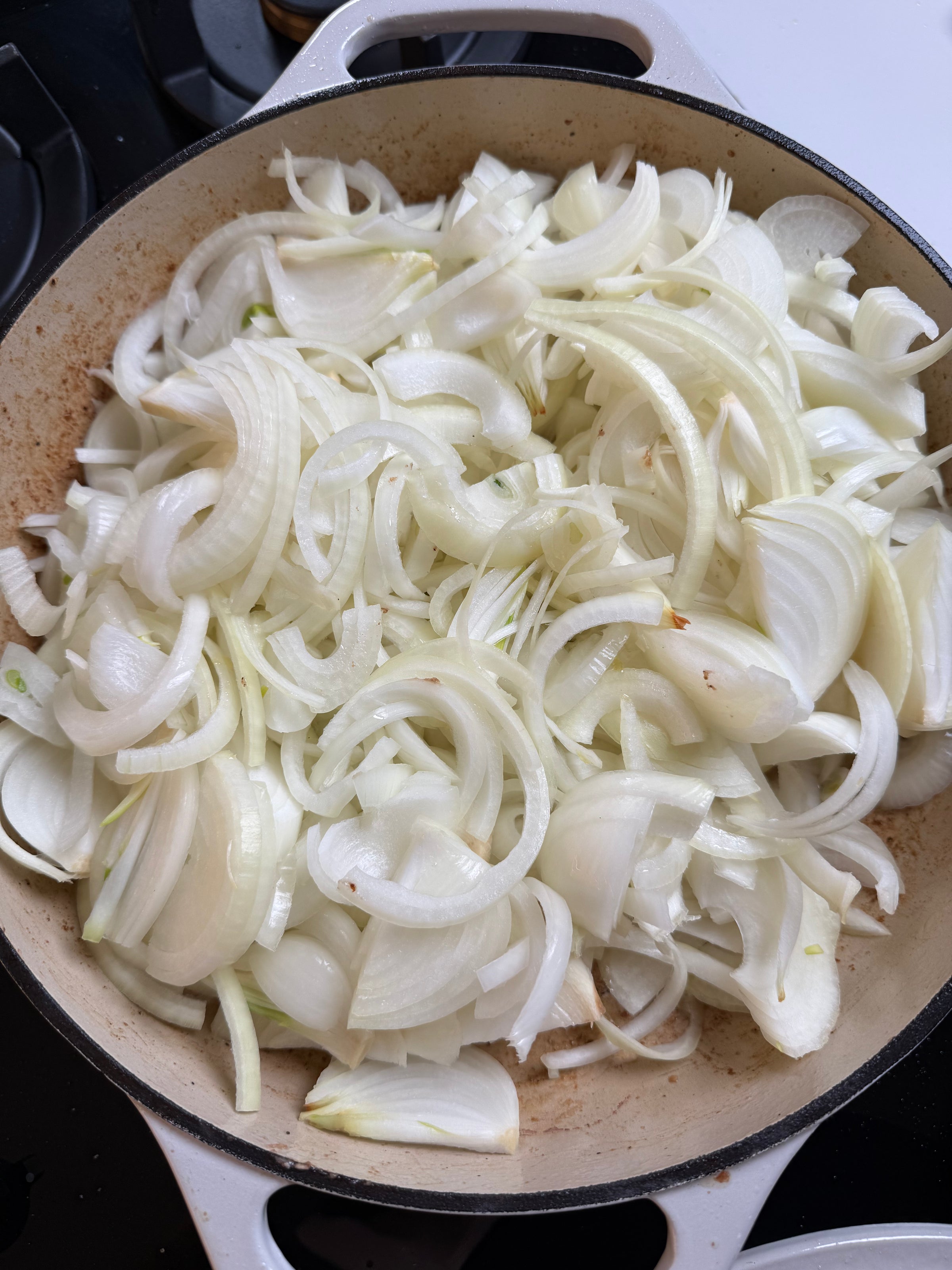 A large white pot filled with a generous amount of thinly sliced white onions, ready to be cooked on a stovetop. The onions are fresh and overlapping in layers.