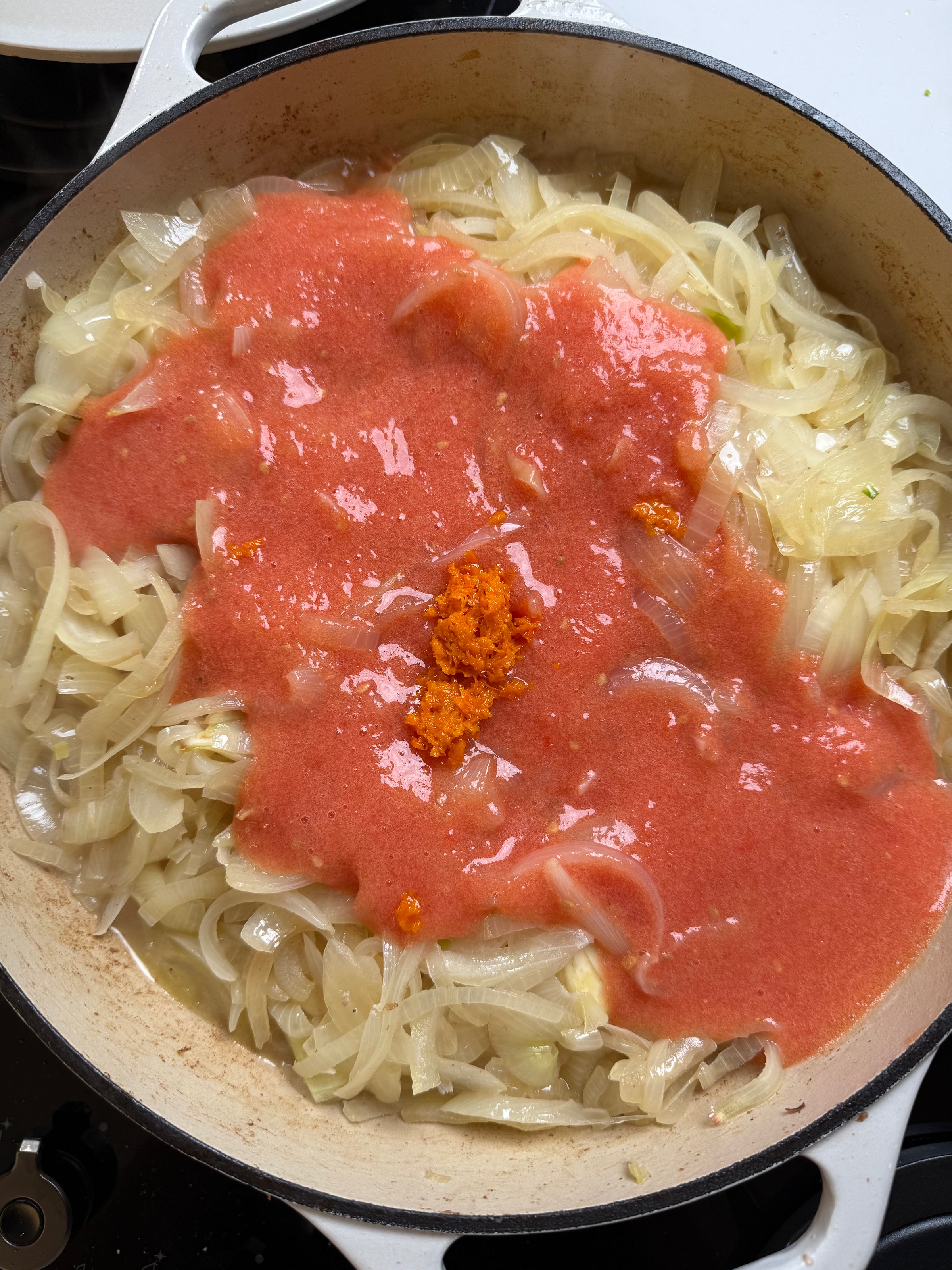A pot of sliced onions being sautéed, topped with a layer of blended tomatoes and a small spoonful of orange-colored paste in the center.