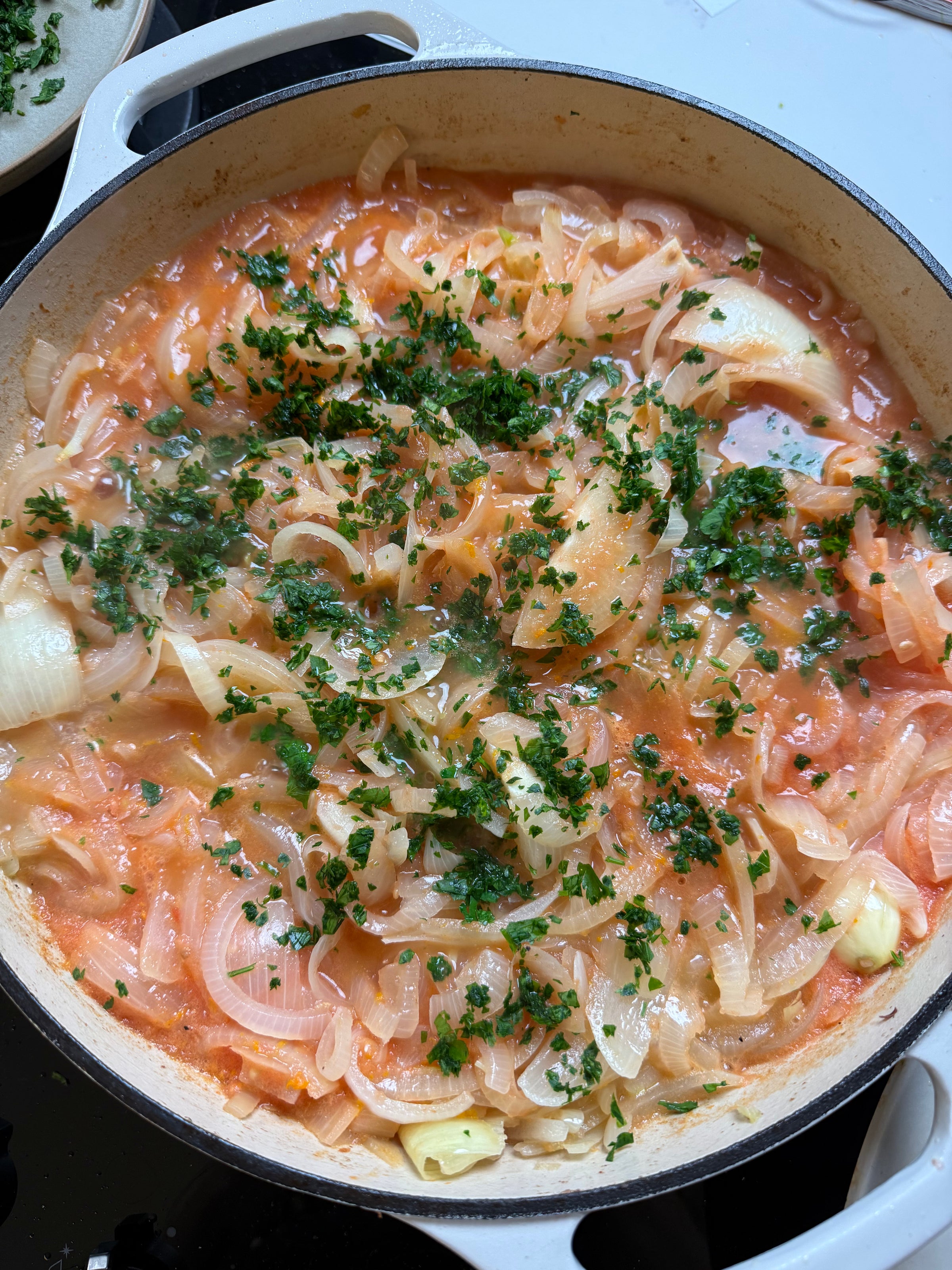 Large white pot on a stove filled with sliced onions, chunks of white fish, tomato sauce, and chopped parsley, all simmering together. Garlic cloves are visible among the ingredients.