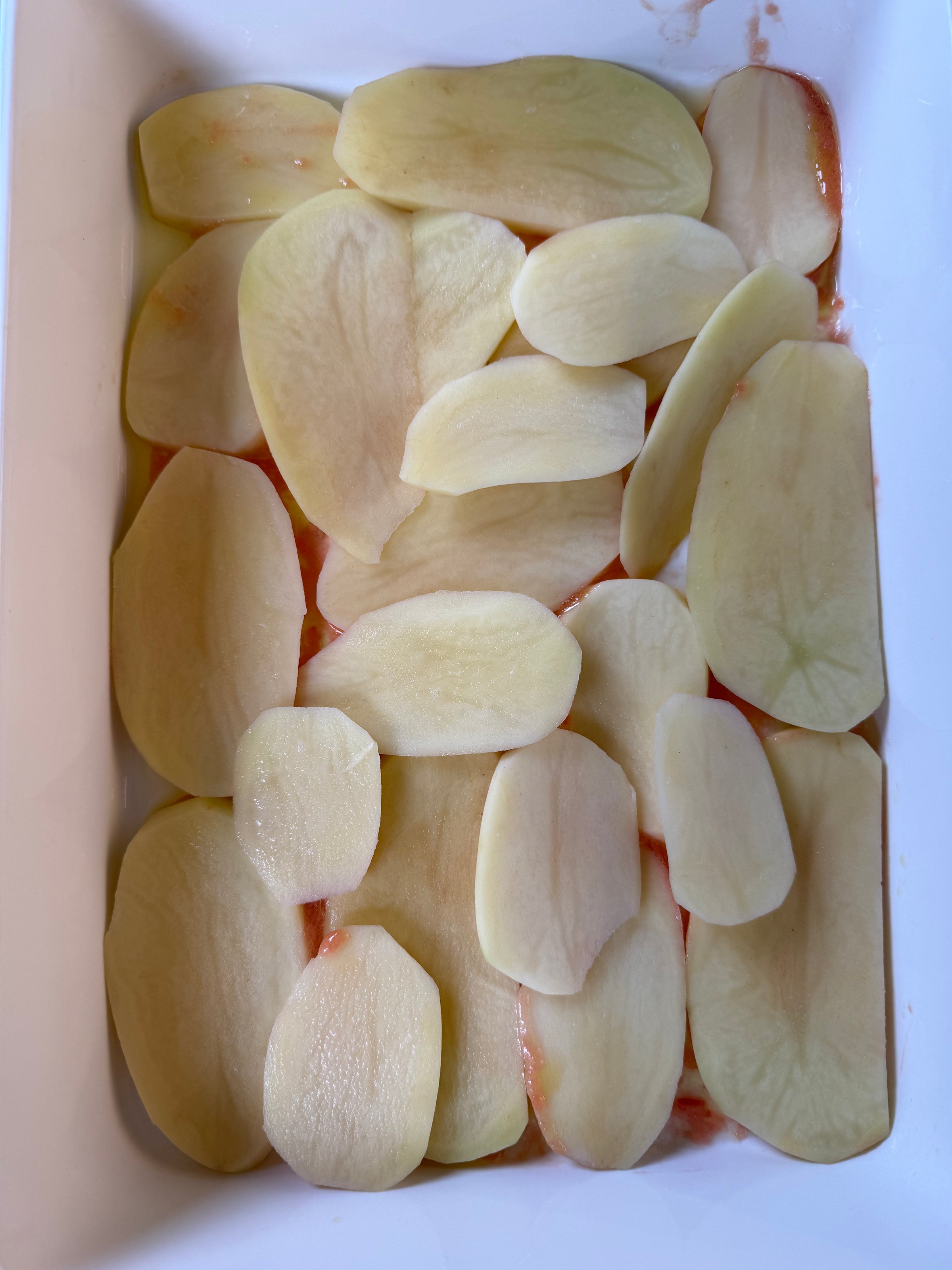 Thinly sliced raw potatoes are arranged in overlapping layers in a white rectangular baking dish. Some tomato sauce is visible underneath the potatoes.