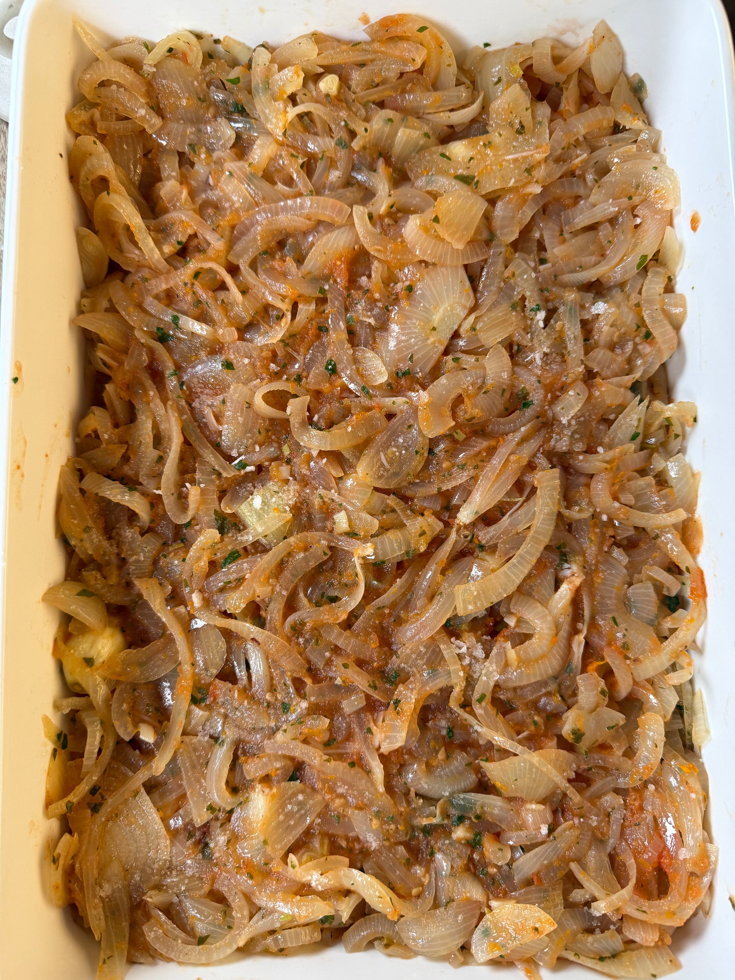 A white baking dish filled with sliced cooked onions mixed with herbs and spices, spread evenly across the surface. The onions appear soft and translucent with a light orange seasoning.