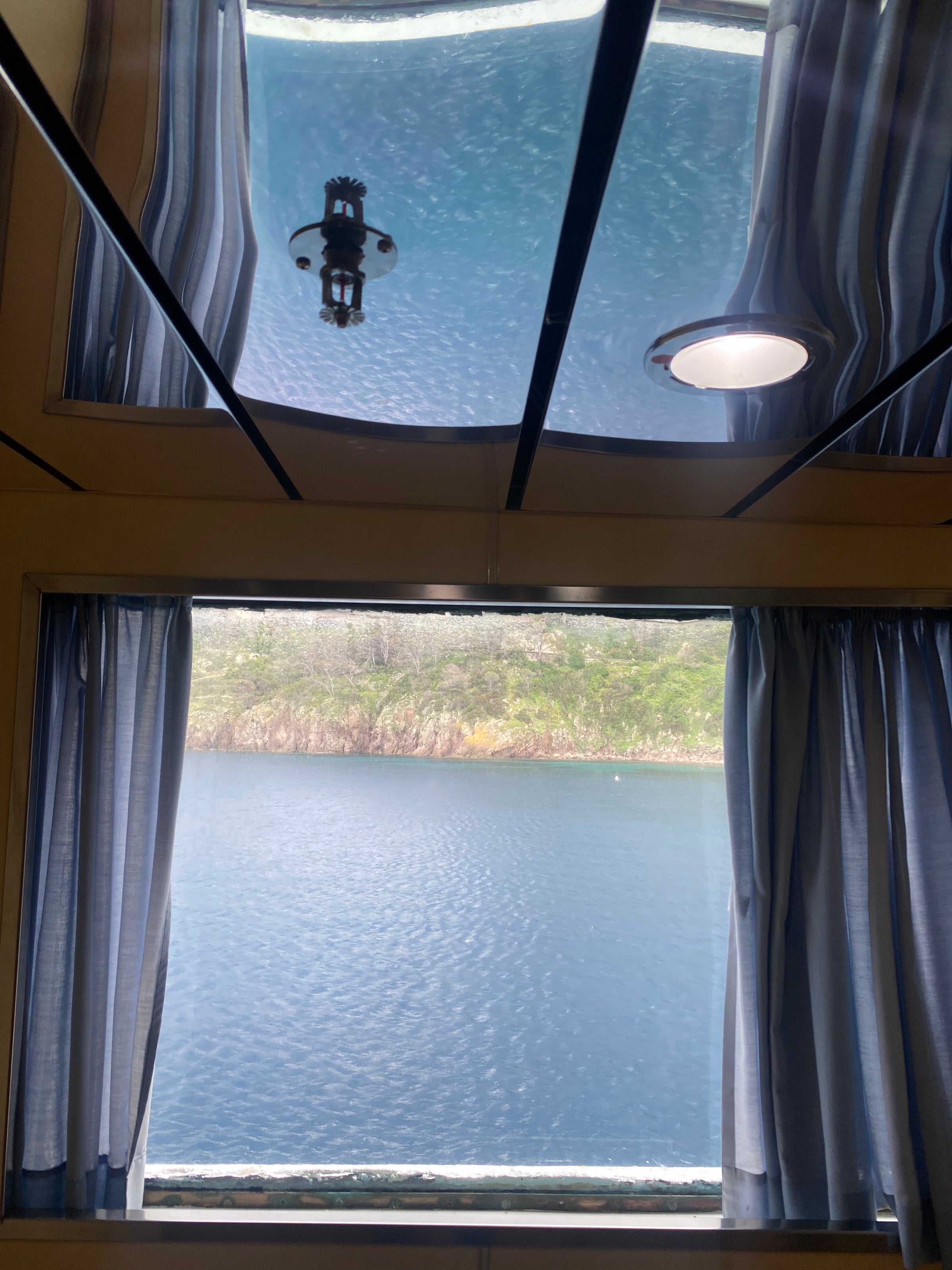 View from inside a room with blue curtains, looking out a window at calm blue water and a grassy, rocky shoreline. The ceiling reflects the water above, showing an underwater scene.