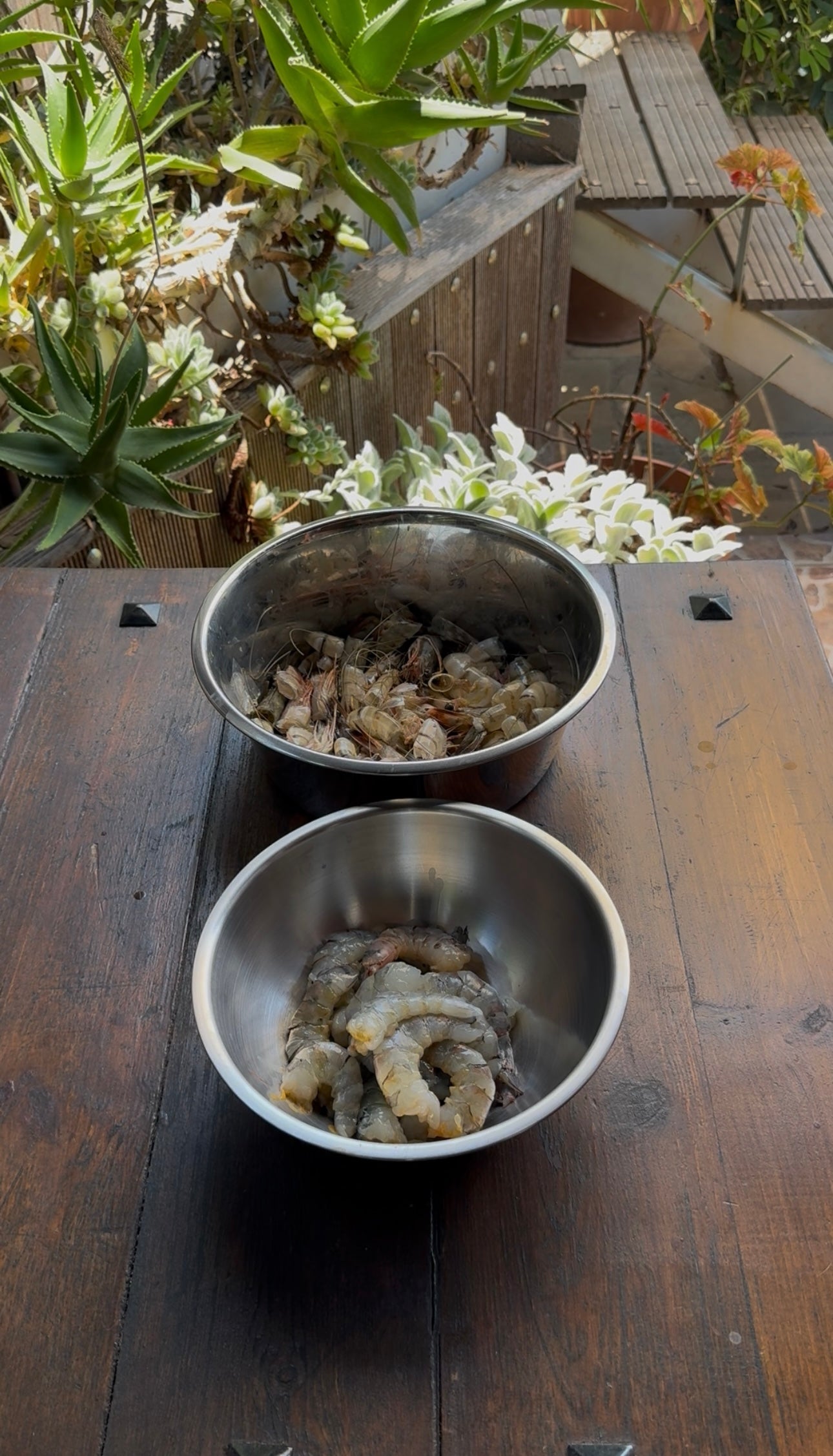 Two metal bowls on a wooden table outside; the front bowl contains peeled shrimp, while the back bowl holds shrimp shells and heads. Green potted plants and a wooden deck are visible in the background.