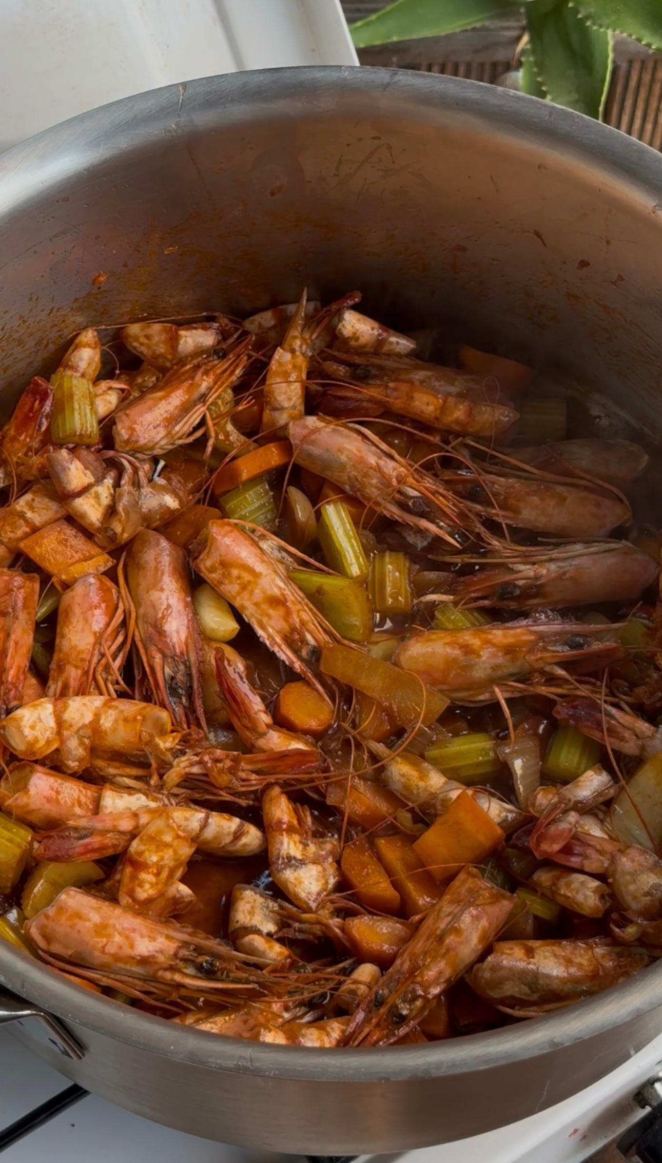 A close-up of a pot filled with shrimp shells, celery, carrots, and onions simmering together, likely to make a seafood stock. The ingredients are coated in a reddish broth.