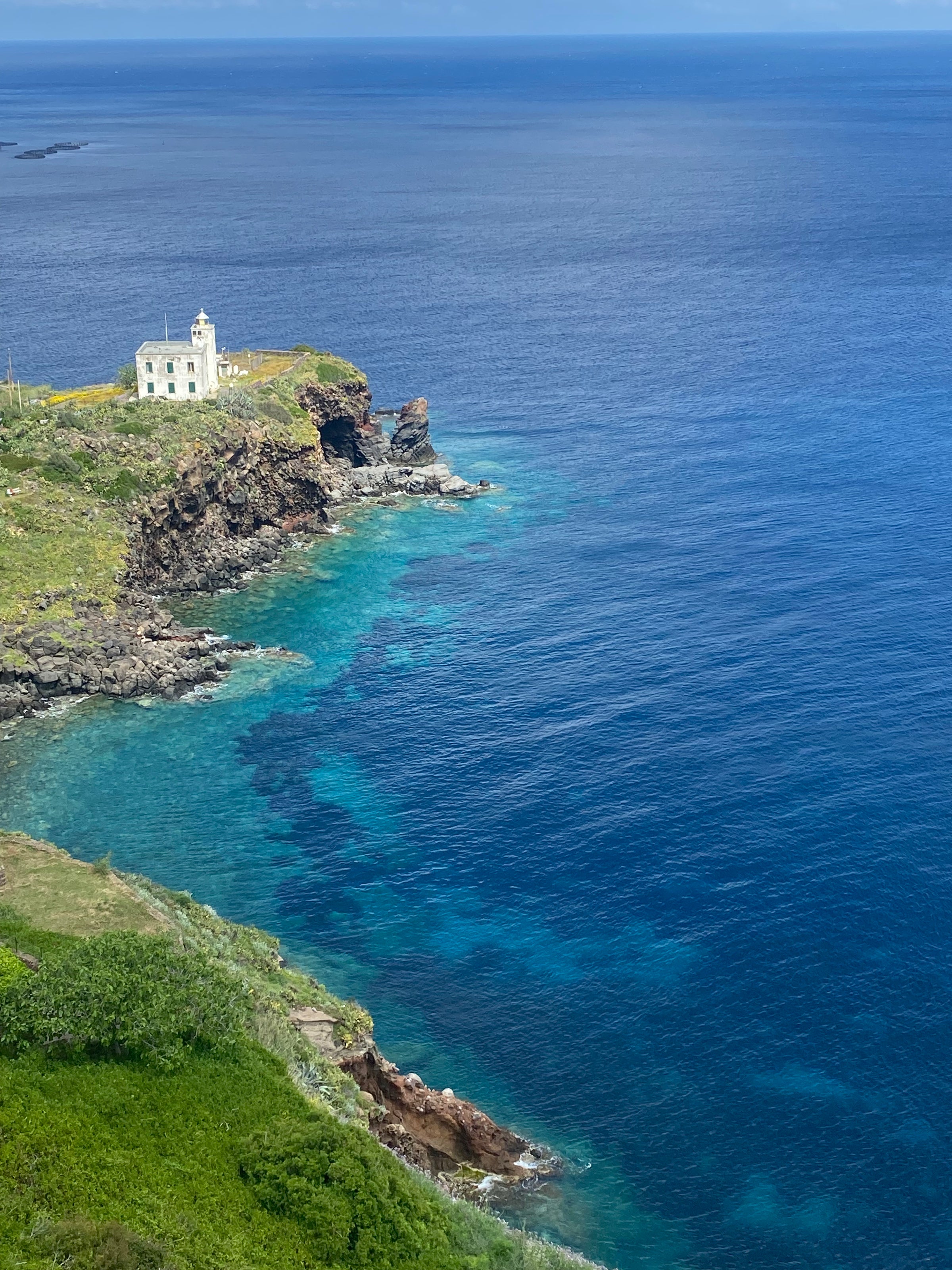 A white lighthouse stands on a rocky green cliff overlooking clear blue ocean water, with deeper blue further from the shore under a sunny sky.