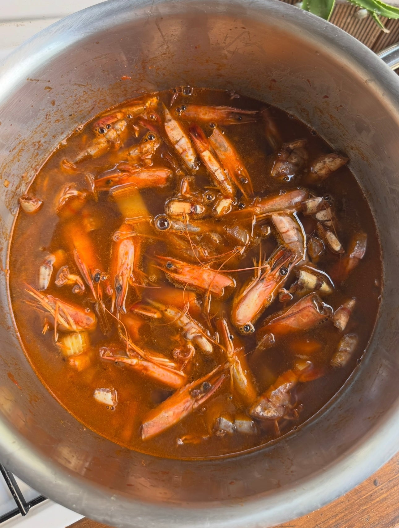 A pot filled with shrimp shells and heads simmering in a reddish broth, likely being cooked to make seafood stock or soup.