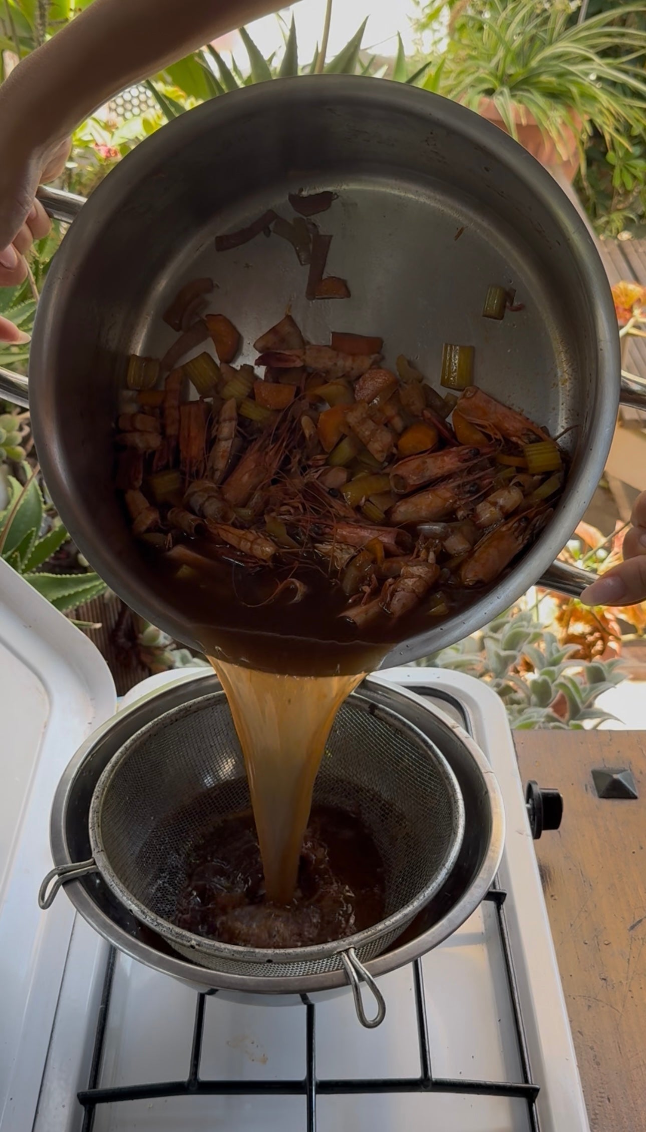 A person pours shrimp shells, vegetables, and broth from a pot through a strainer over a bowl outdoors, surrounded by lush green plants.