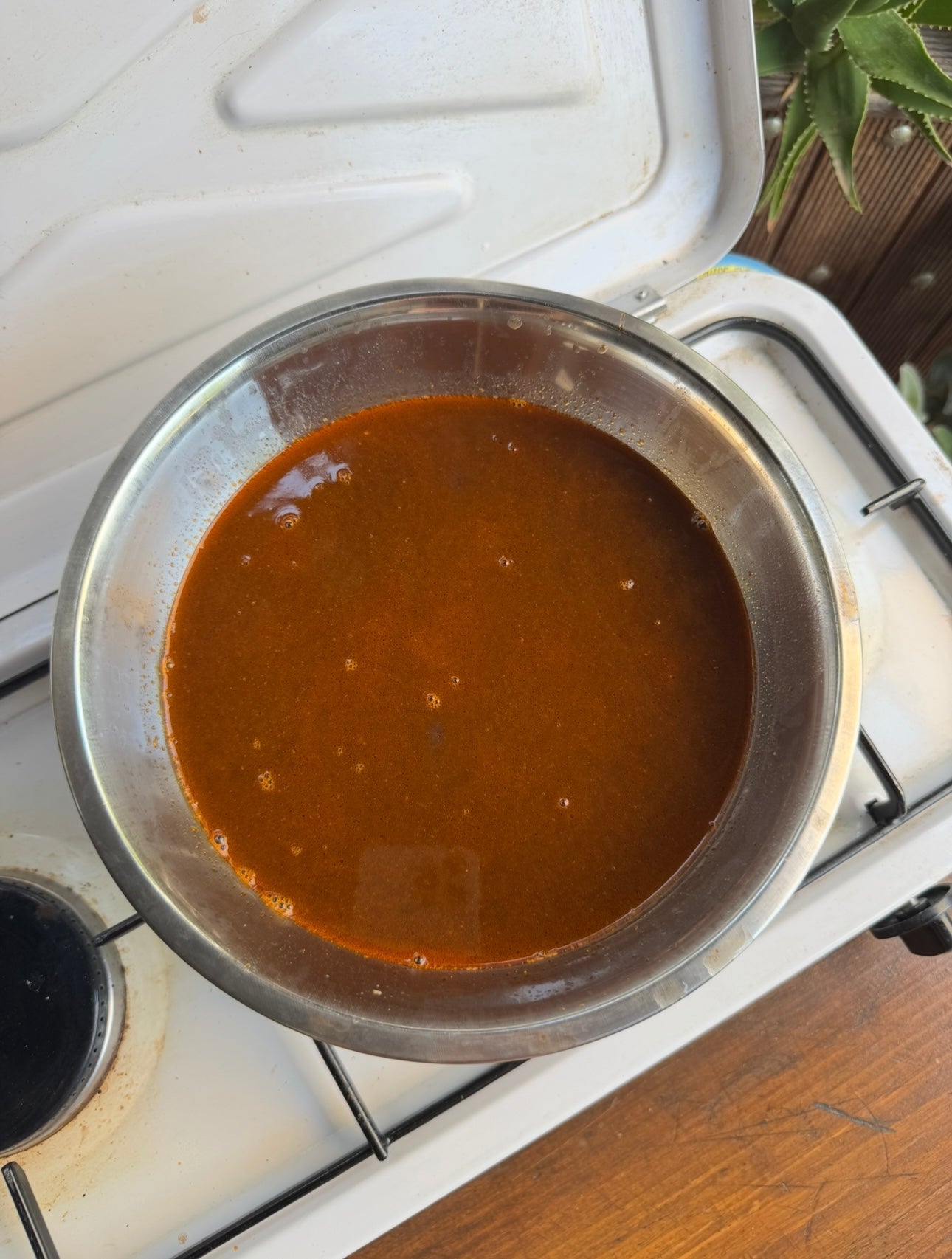 A metal pot filled with reddish-brown sauce sits on a white gas stove, with bubbles visible on the sauce’s surface. A plant and a wooden surface are partly visible in the background.