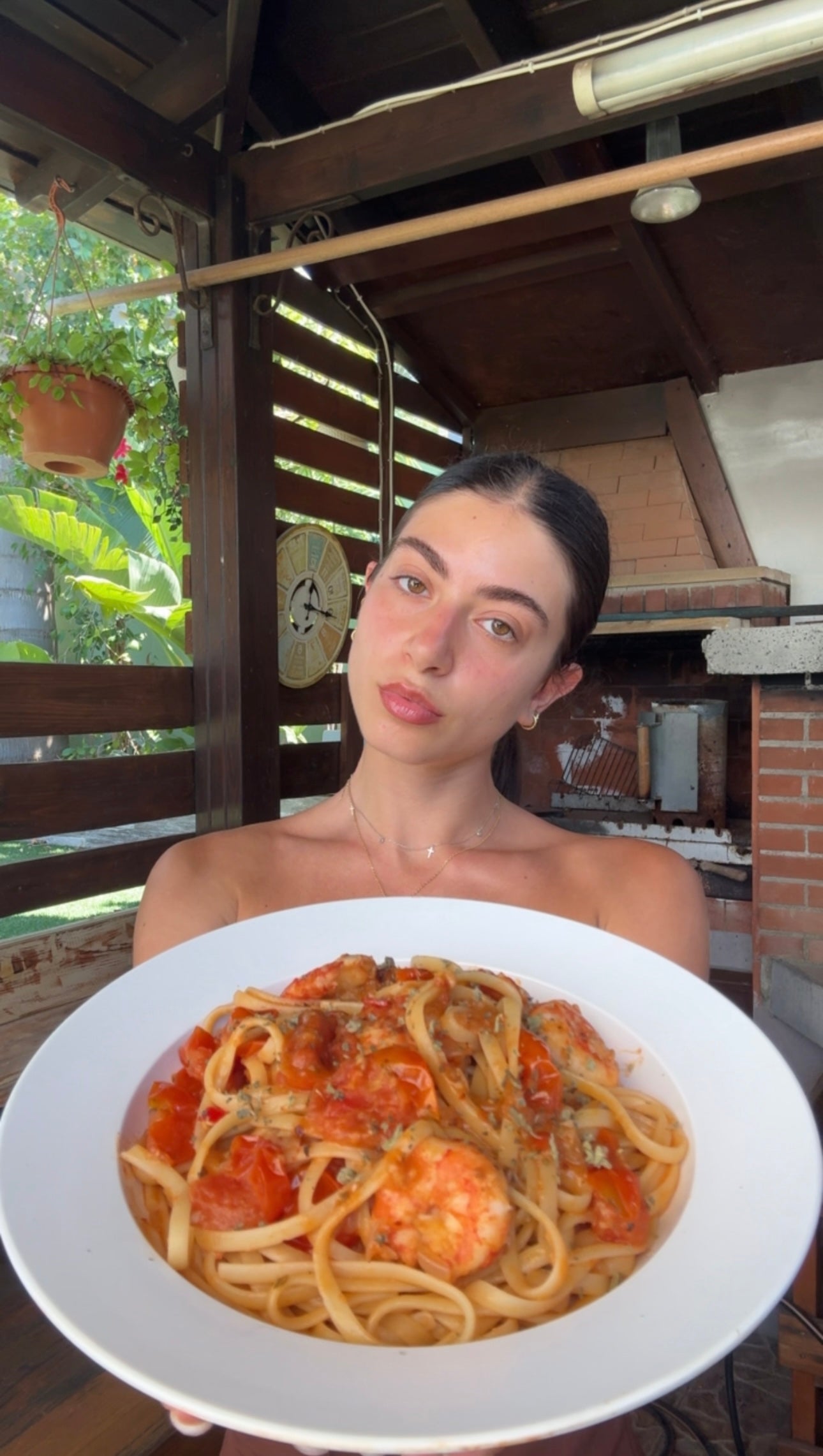A woman with dark hair pulled back poses in an outdoor kitchen, holding a large white bowl of pasta with shrimp, tomato sauce, and herbs. There are plants and a brick oven in the background.