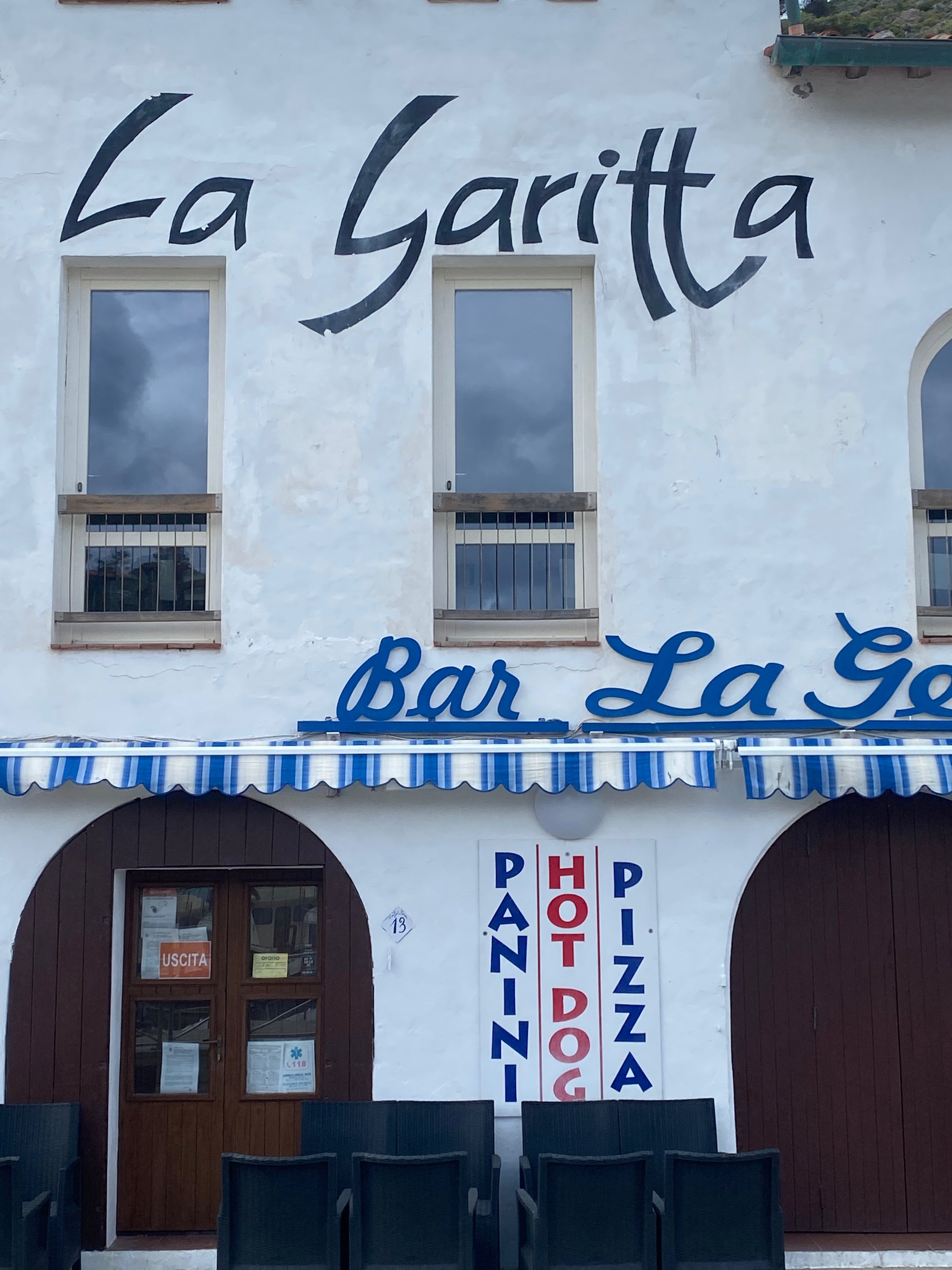 A white building with La Garitta painted on it, blue-striped awning, and a sign listing Panini, Hot Dog, Pizza vertically beside a wooden door. Several empty black chairs are visible in the foreground.
